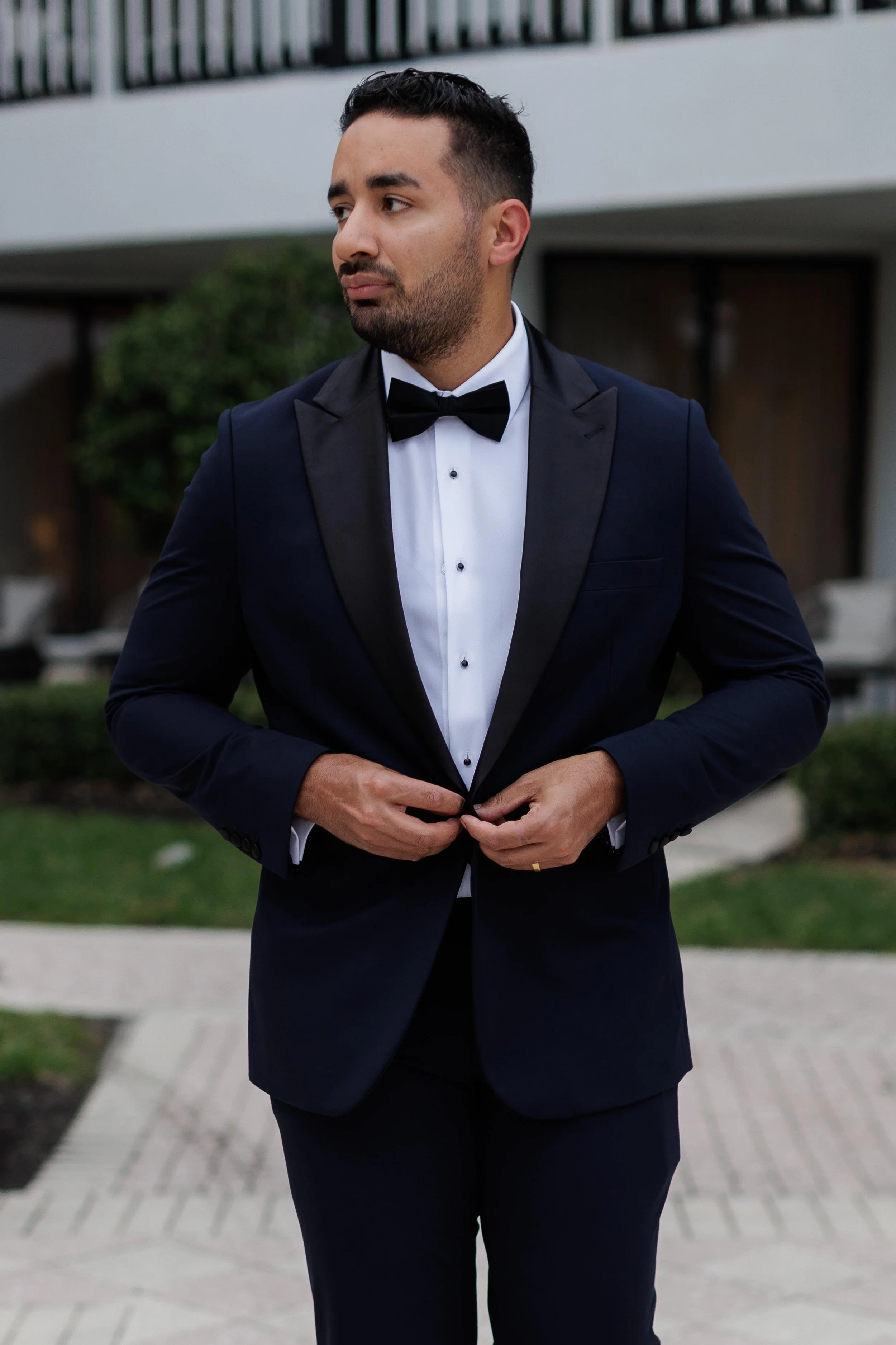 A man in a tuxedo buttoning his jacket outdoors, with modern building and greenery in the background.