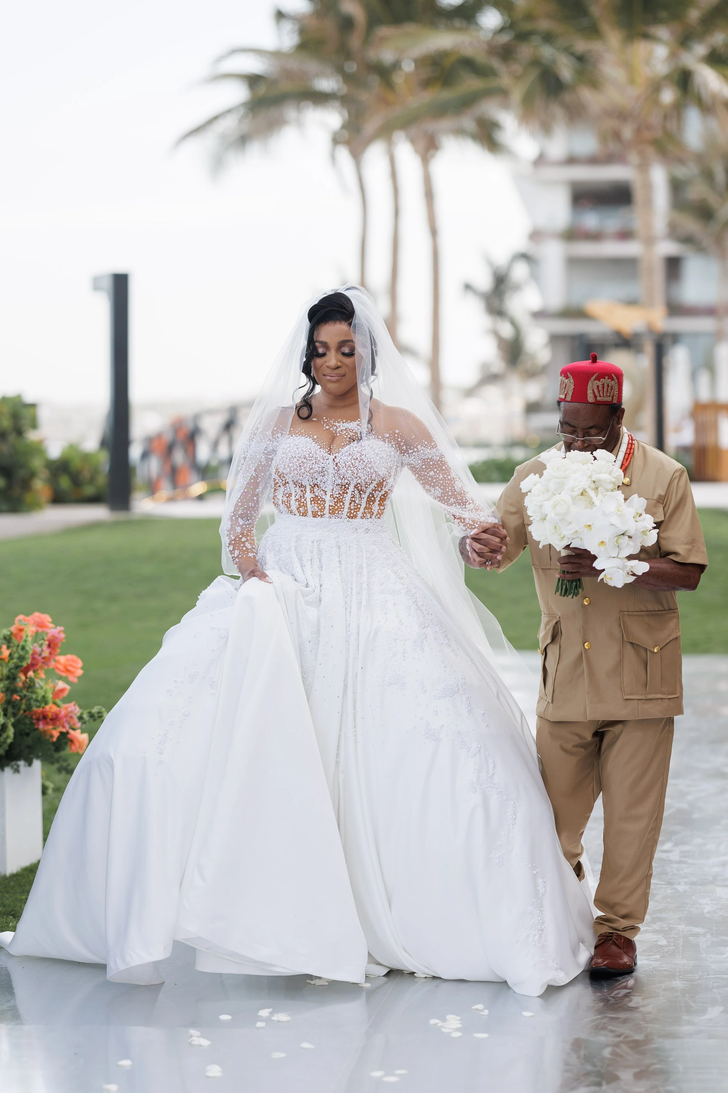 A bride in a white wedding dress holding hands with a man in traditional attire, walking outdoors on a green lawn with palm trees and buildings in the background.