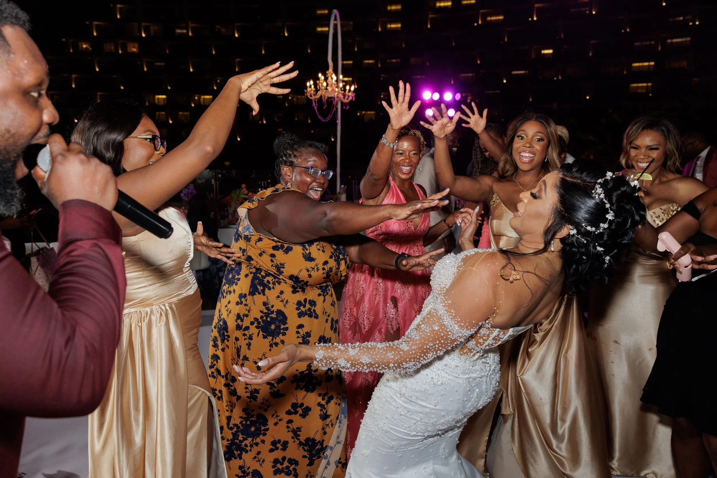 People dancing and celebrating at a wedding reception, with a woman in a white wedding gown in the center and others smiling and reaching toward her.