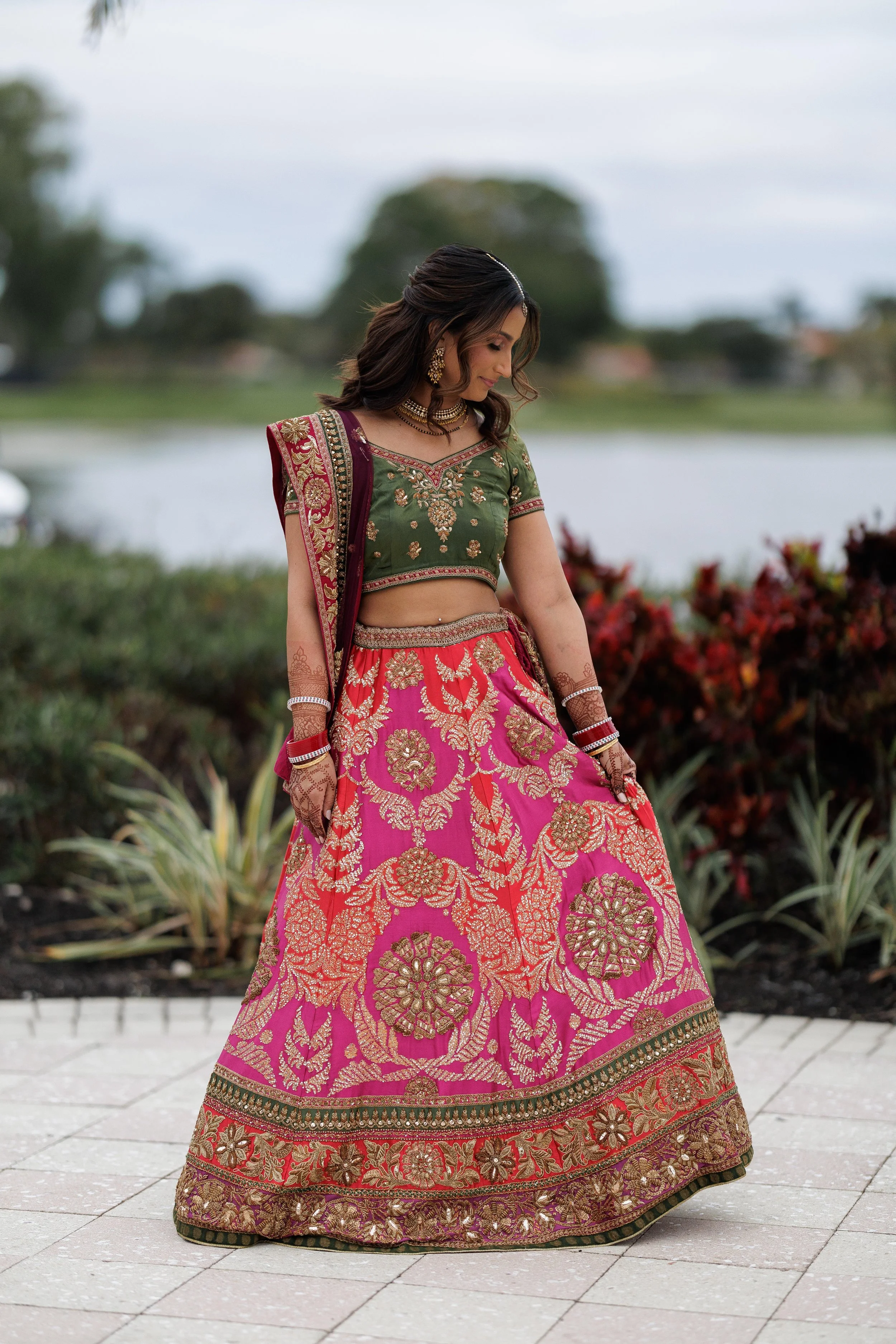 A woman wearing a traditional Indian outfit, consisting of a green embroidered blouse and a pink and gold embroidered skirt, standing outdoors near a lake or river, with trees and clouds in the background.