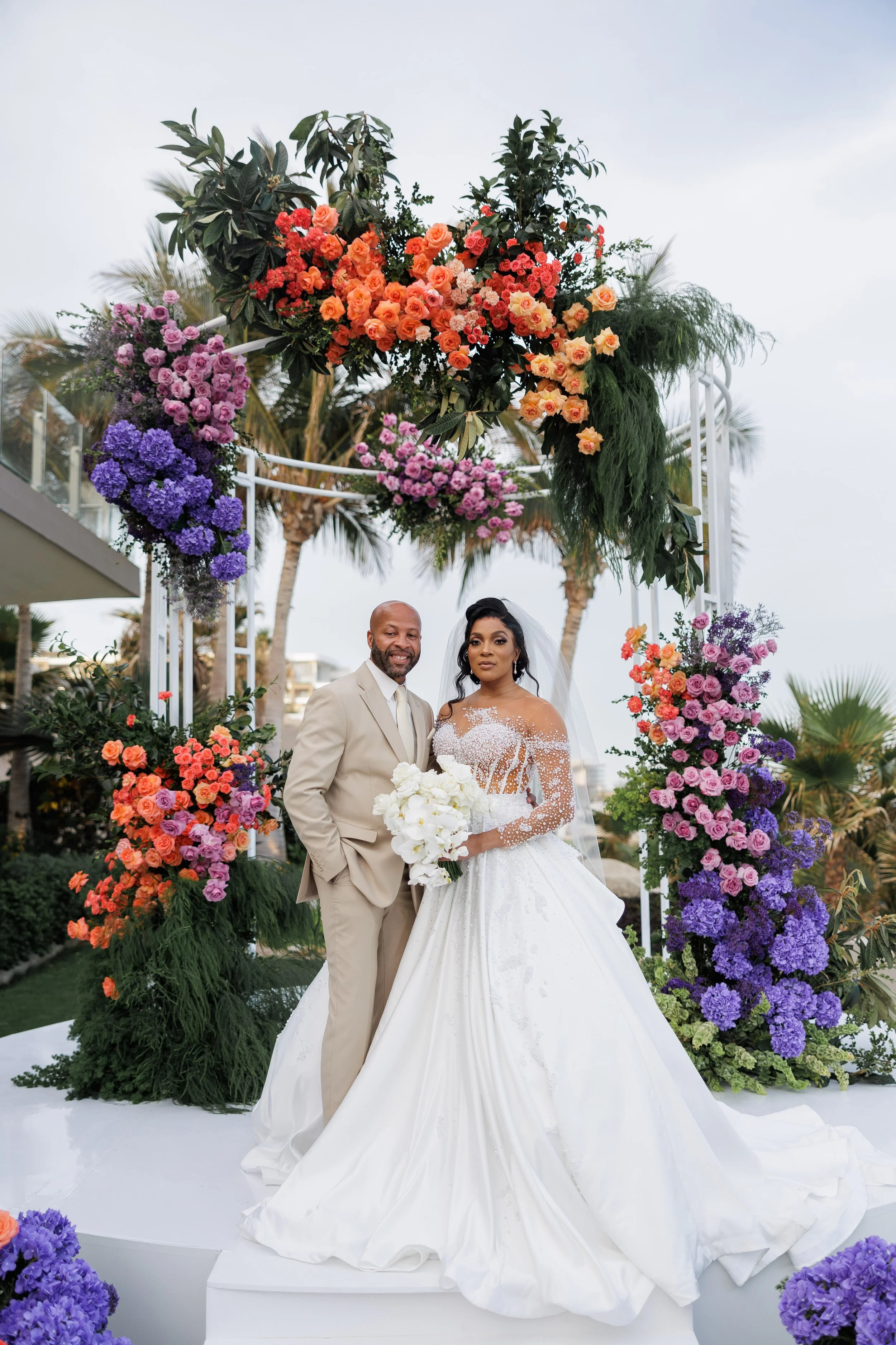 A newlywed couple in wedding attire standing under a floral arch on an outdoor stage, surrounded by vibrant purple, pink, and orange flowers and lush greenery, with palm trees in the background.