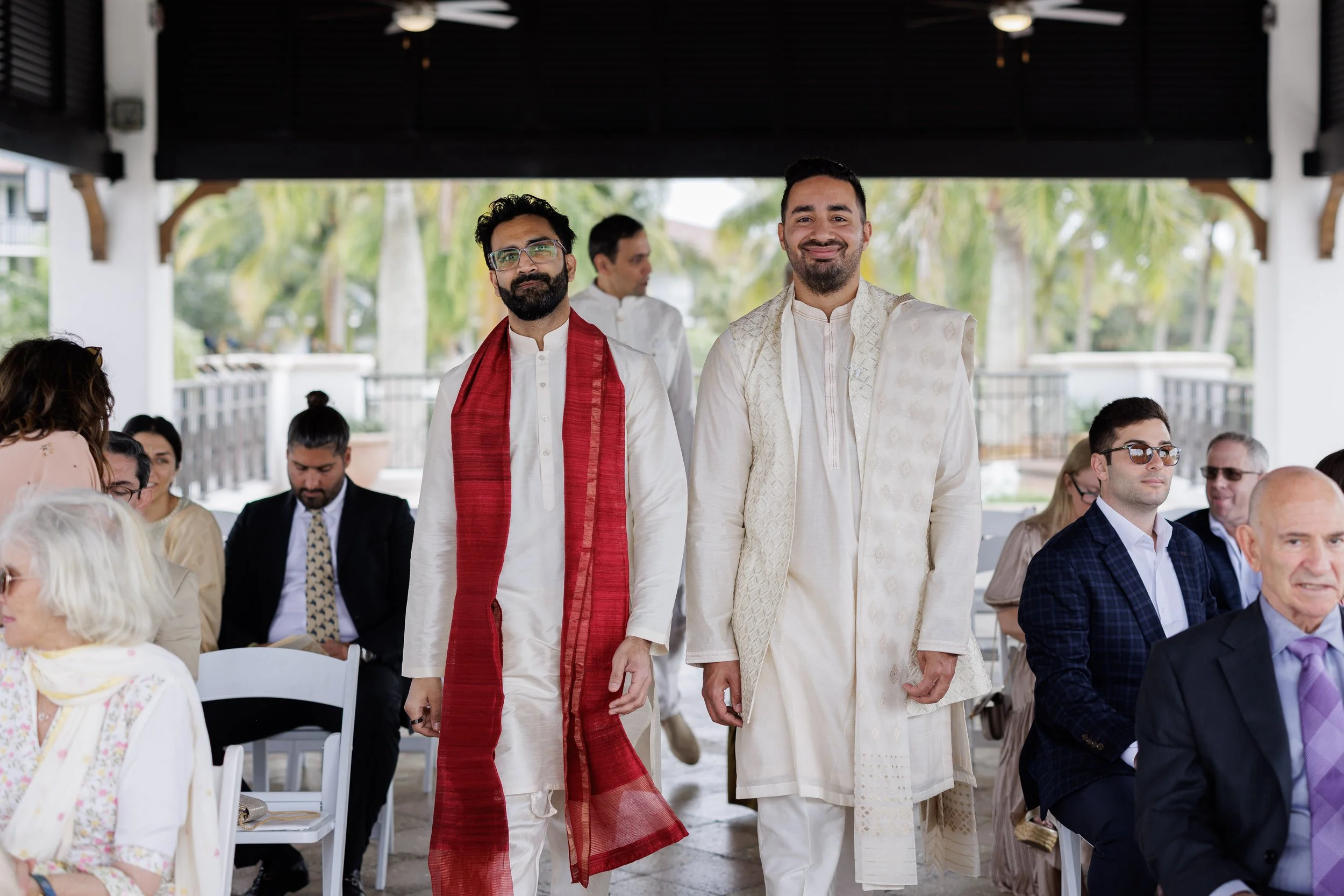 Two men in traditional Indian attire walking at an outdoor wedding ceremony with guests seated at tables around them.