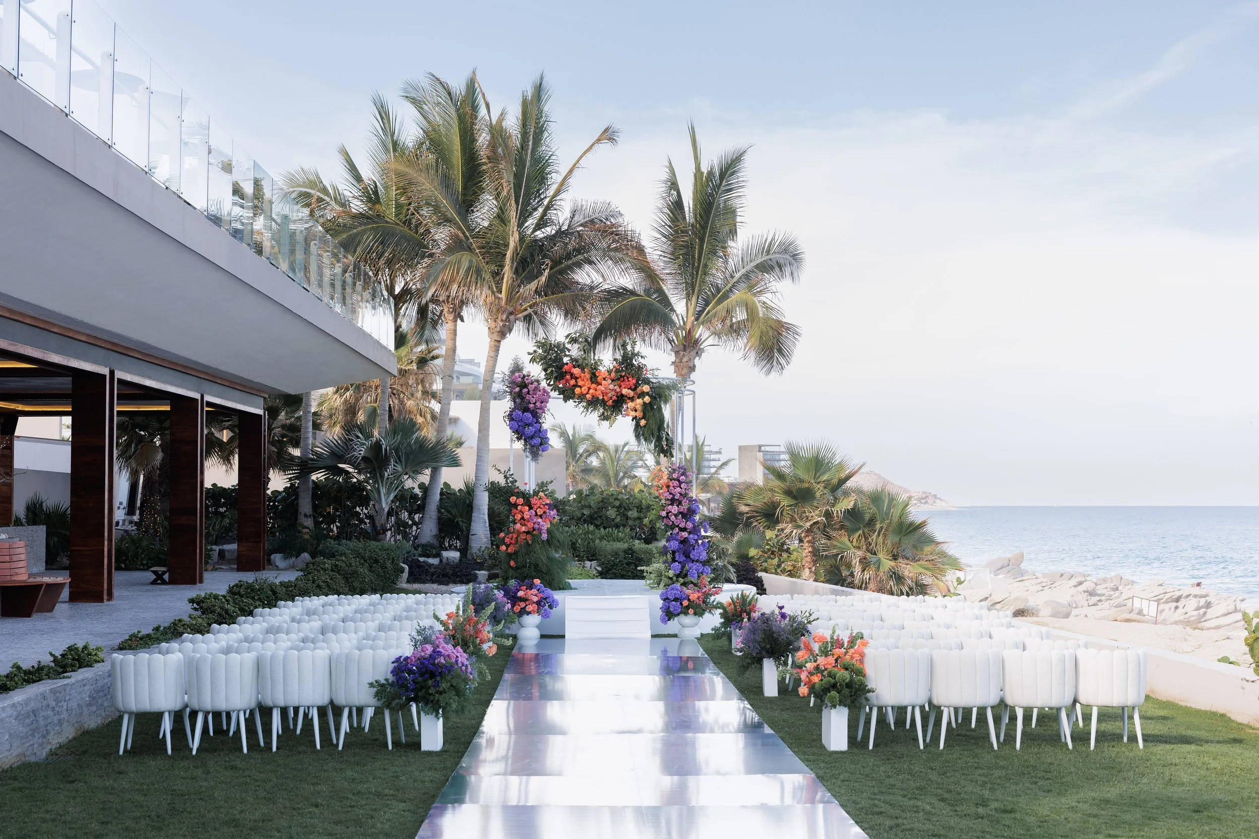 Beachside wedding setup with a white aisle, flower arrangements, and tropical palm trees under a clear blue sky.