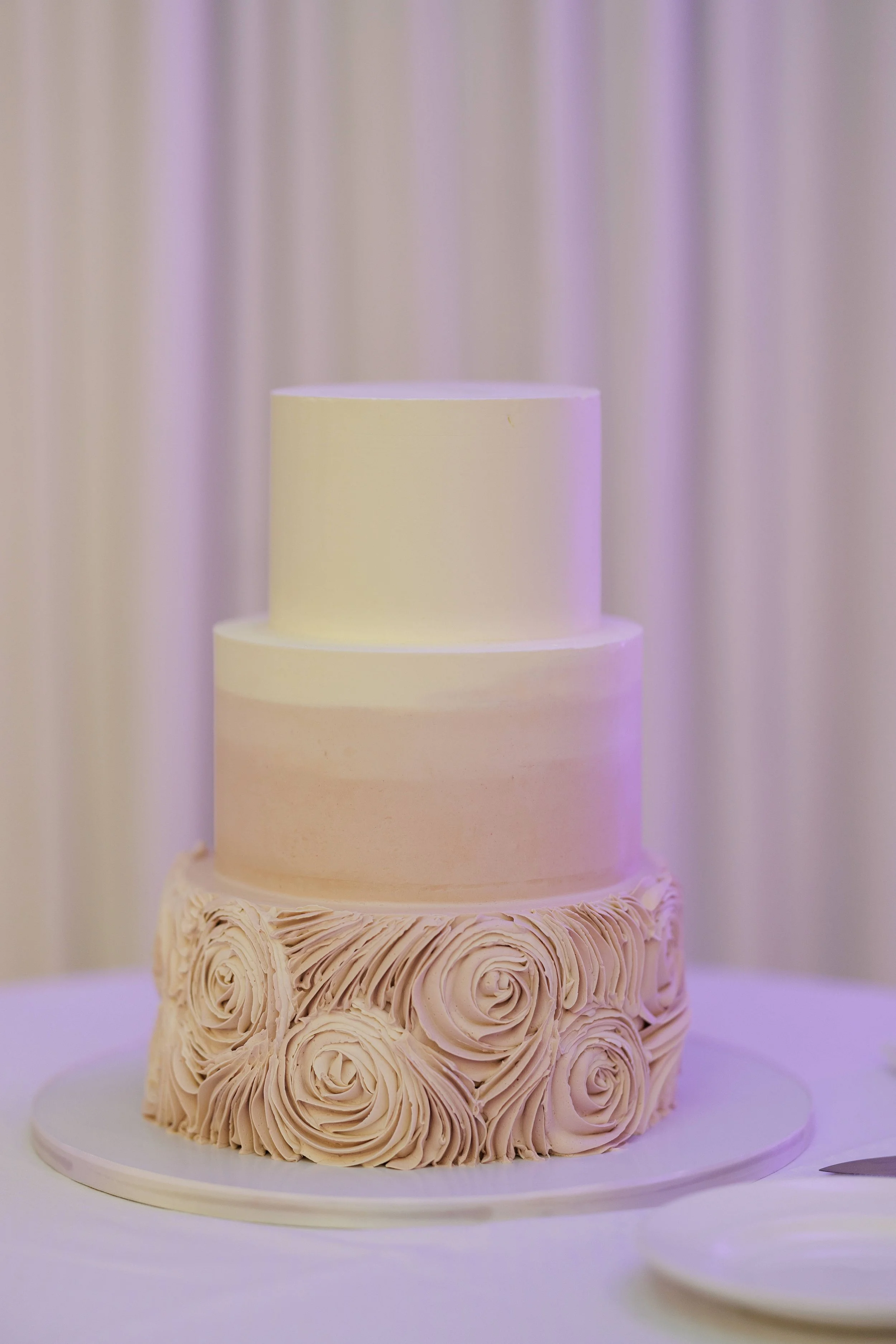 Three-tiered wedding cake with cream-colored rosette decorations at the base, smooth middle, and simple top, on a white table with a white plate and cake knife nearby.