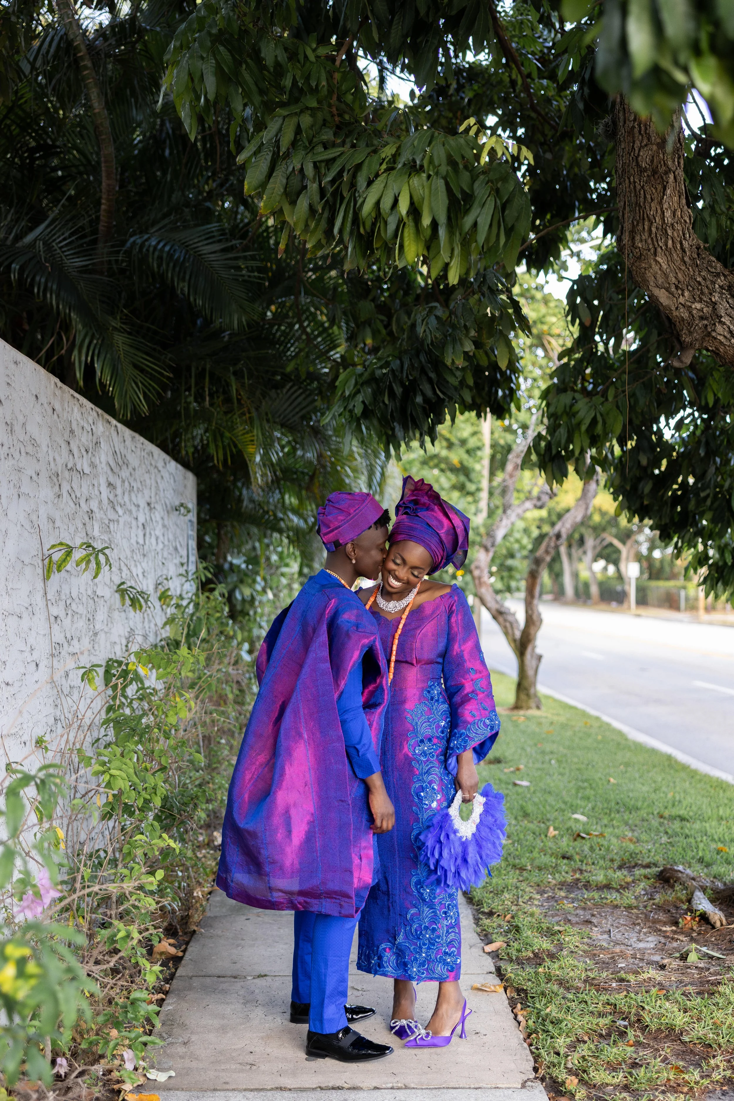 Two women dressed in matching traditional African outfits standing close and smiling under a tree on a sidewalk.
