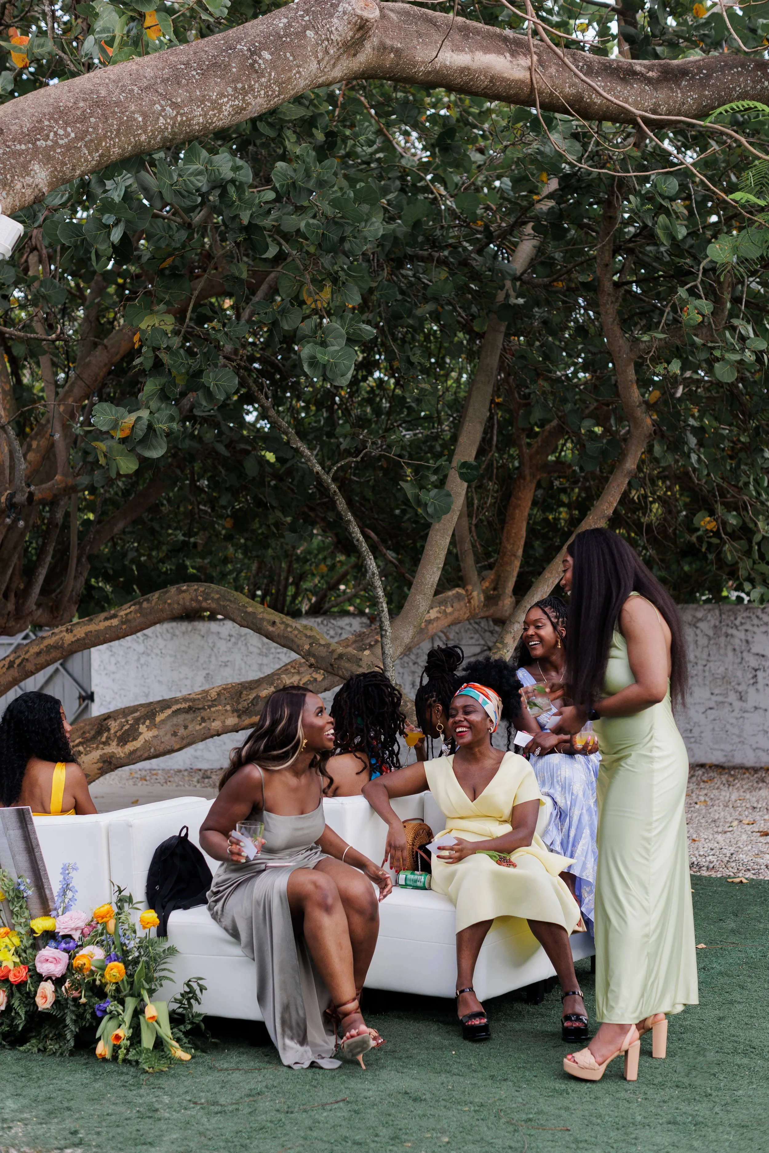 A group of women socializing outdoors under a large tree, sitting on a white couch and a few standing. They are laughing and holding drinks, dressed in colorful dresses with flowers and greenery around.