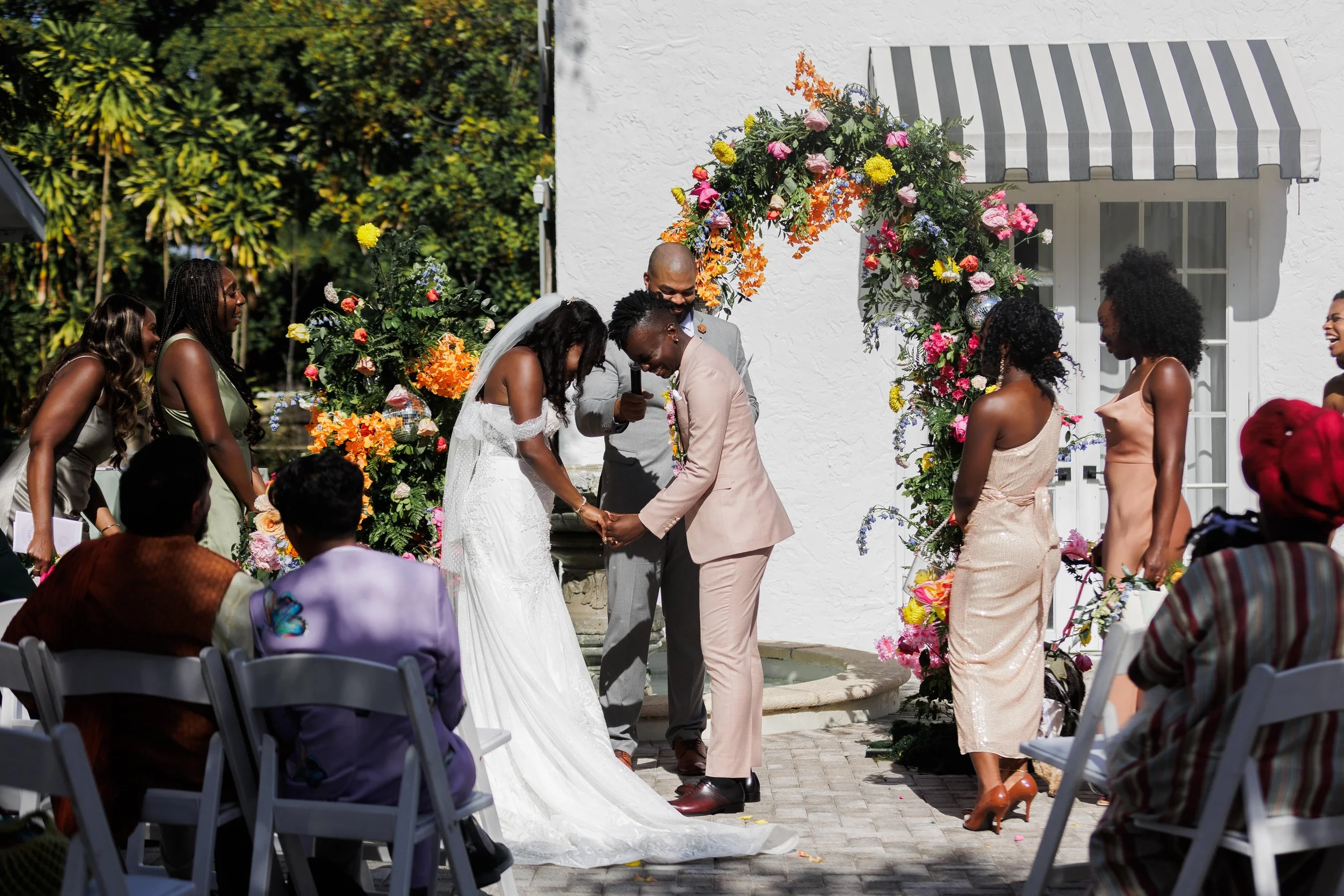 A wedding ceremony outdoors with a bride and groom holding hands, surrounded by bridesmaids, groomsmen, and guests under a floral arch, on a sunny day.