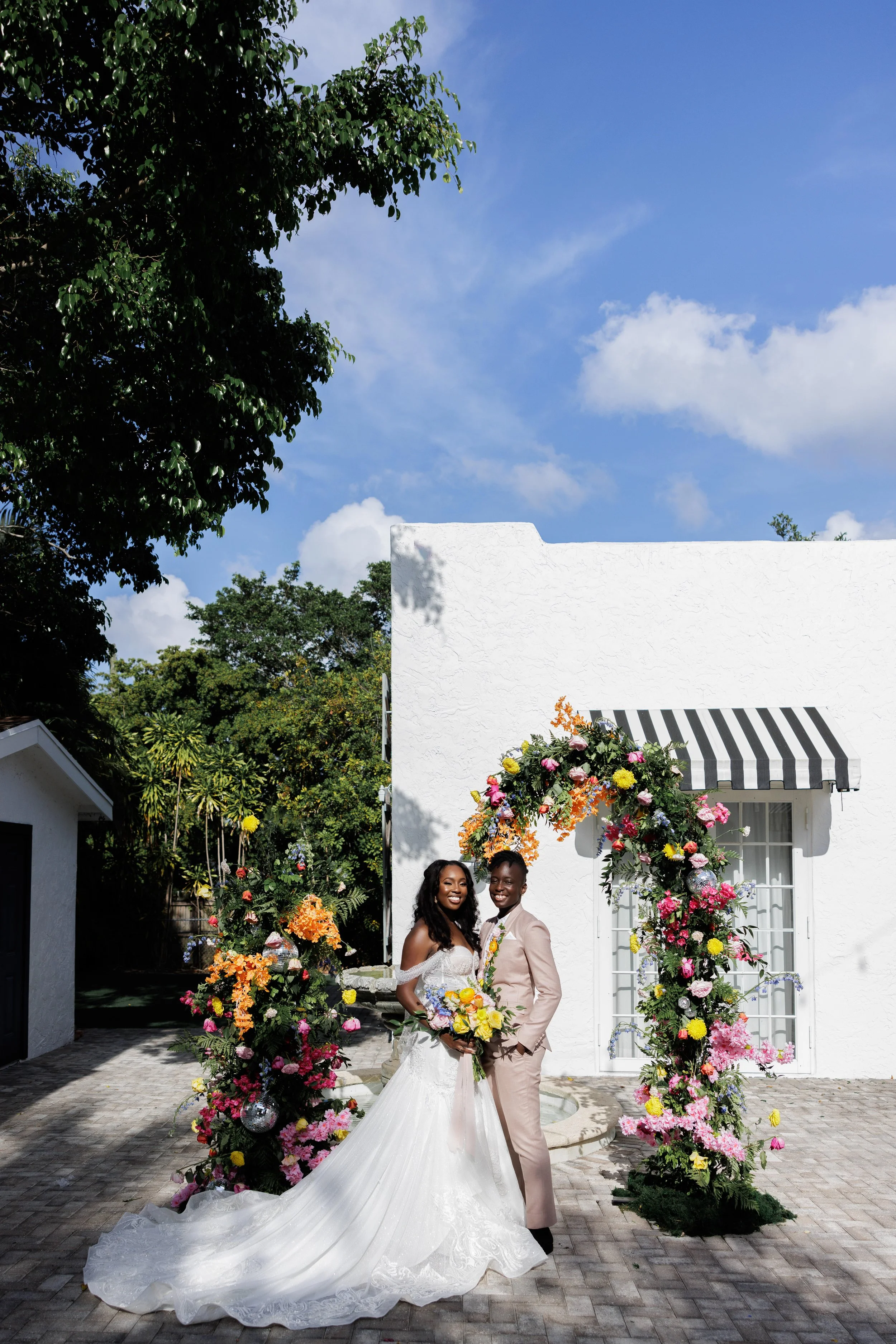 A newlywed couple standing under a colorful flower arch outside a white building with striped awning on a sunny day.