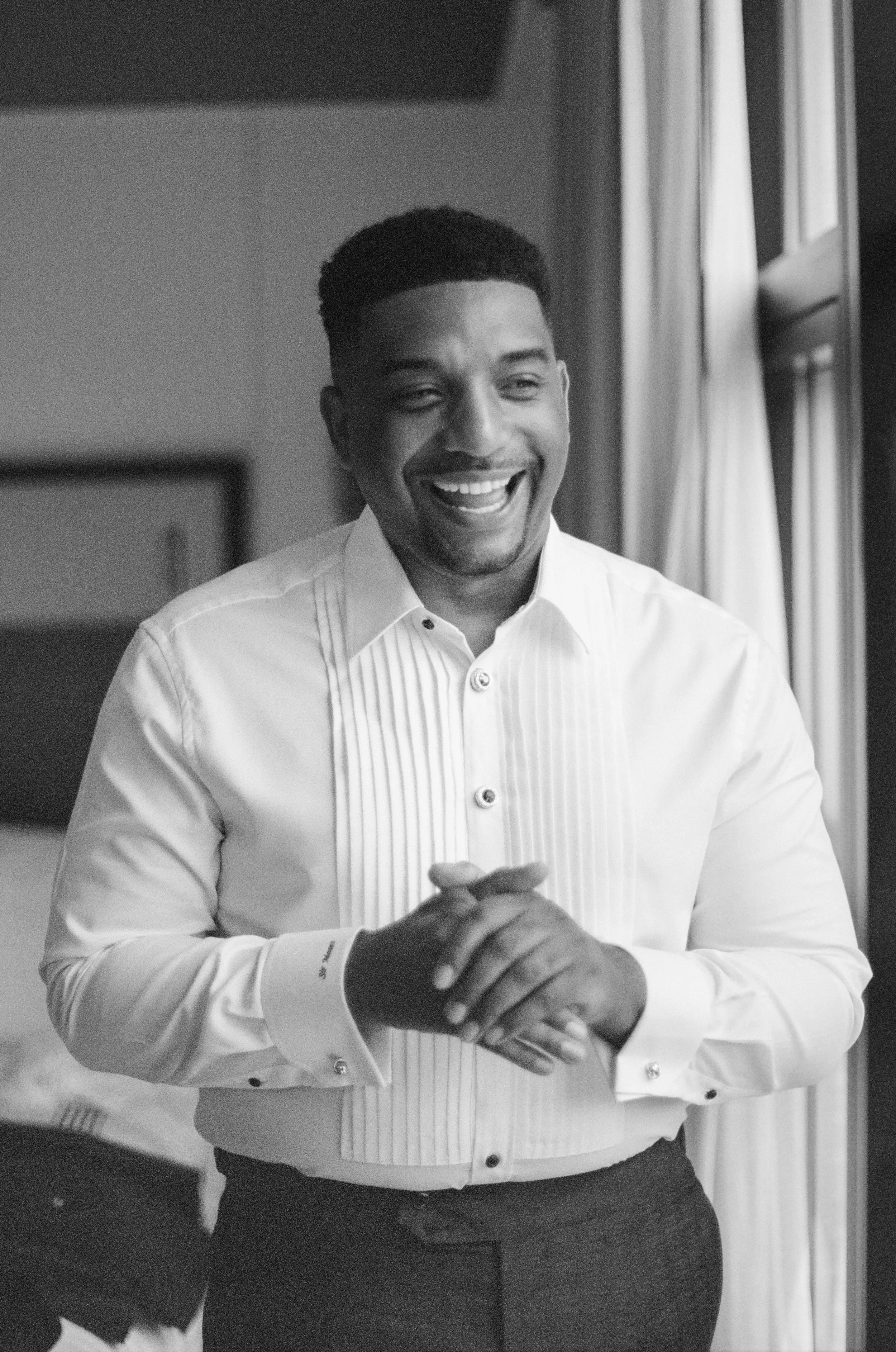 A smiling man in a white dress shirt with pleats, standing near a window with curtains, in a well-lit room.