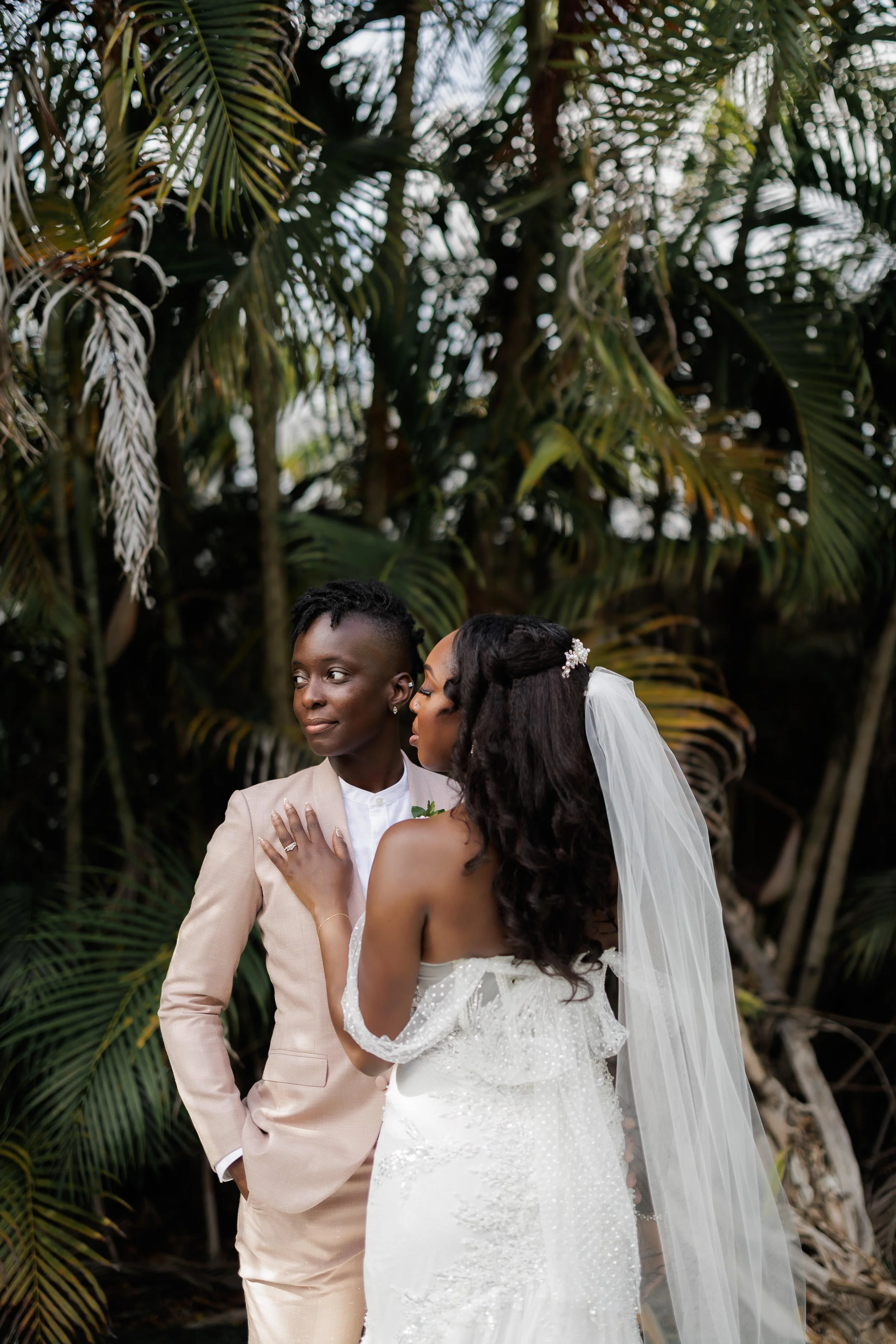 A same-sex wedding photo featuring two women, one in a beige suit and the other in a white wedding dress and veil, standing close together outdoors with tropical palm trees in the background.