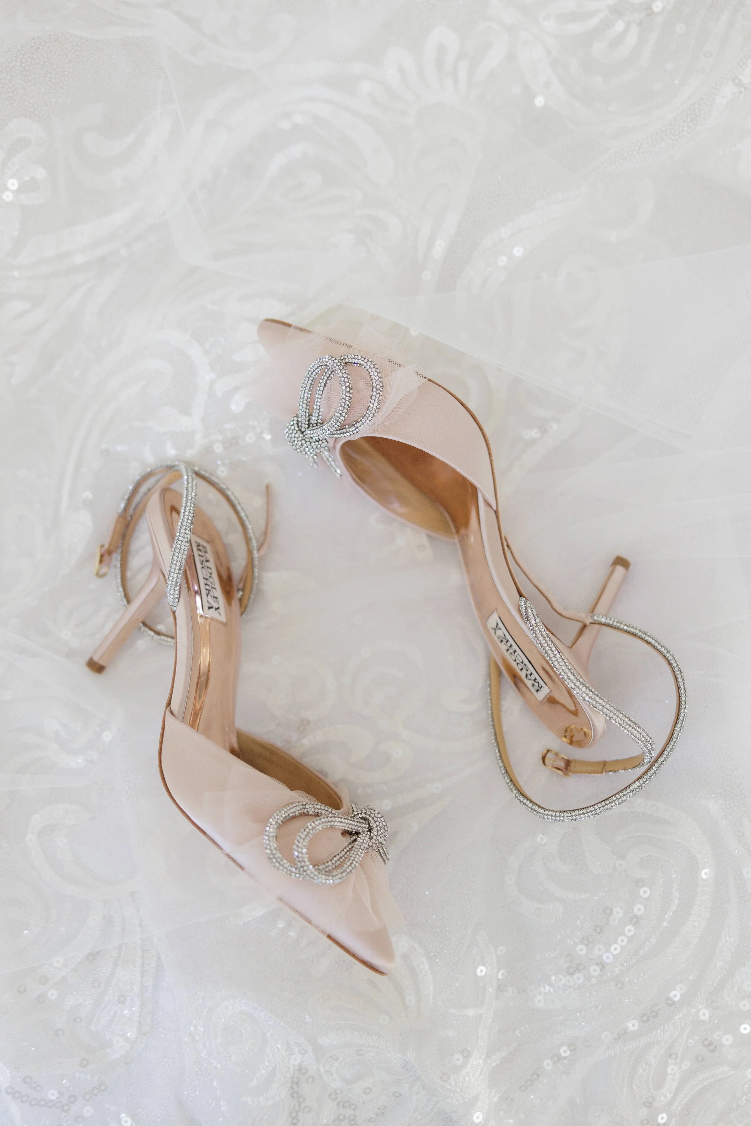 A pair of nude high-heeled shoes with rhinestone embellishments and bows, resting on a white, ornate fabric surface.