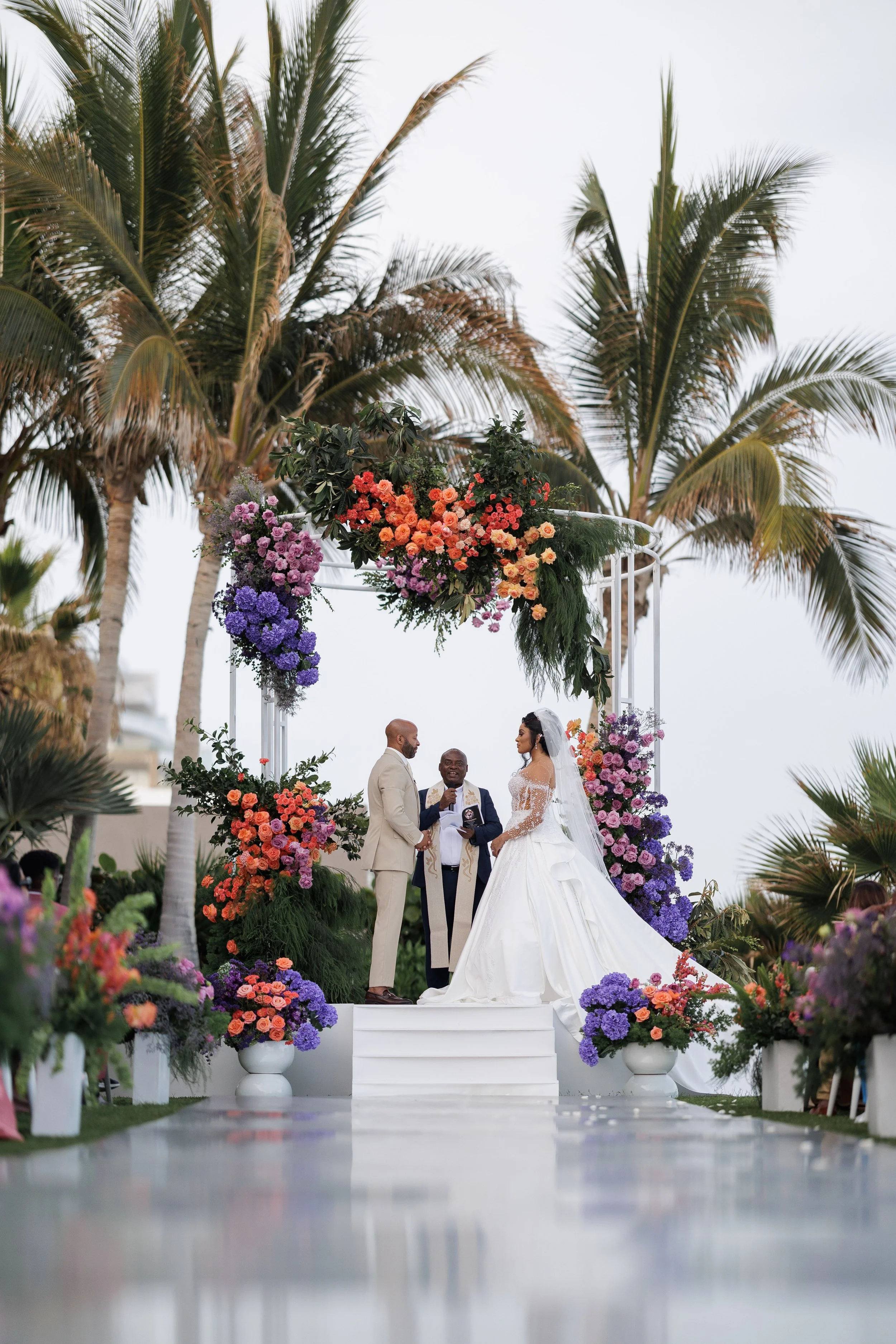 A wedding ceremony outdoors under palm trees with a floral arch, bride in a white wedding dress and groom in a beige suit standing before an officiant.