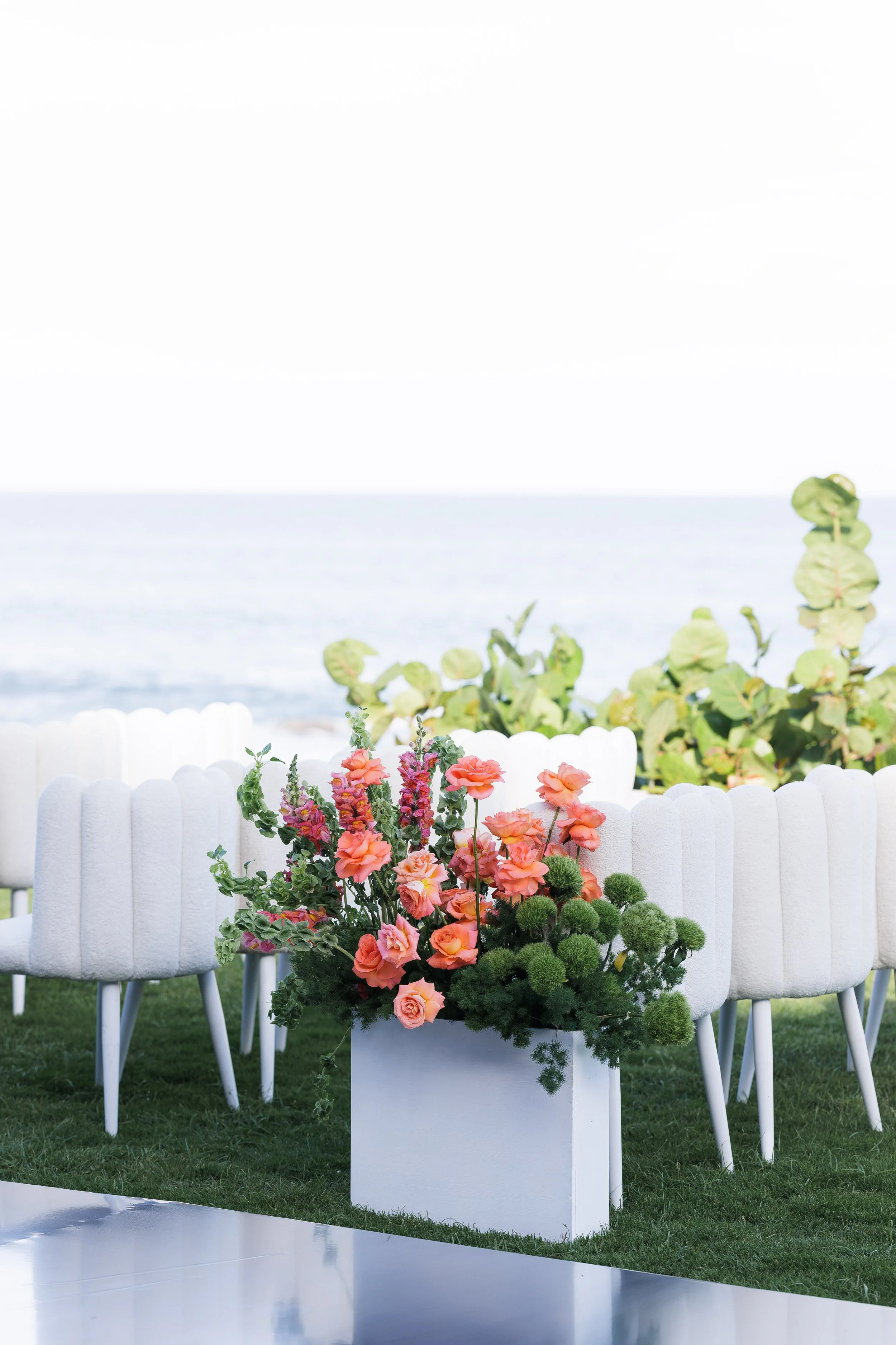 A large white flower arrangement with pink peonies and other flowers in a white rectangular container, set outdoors on green grass with white chairs and green foliage in the background near the beach.