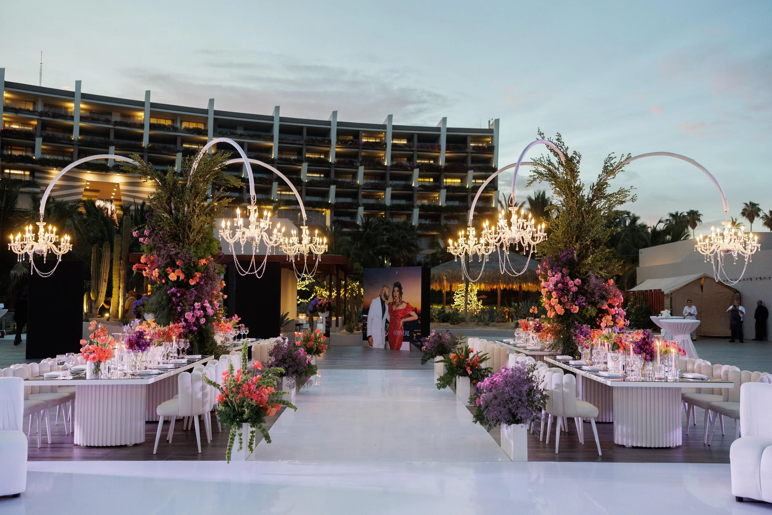 Elegant outdoor event setup with long tables decorated with pink, purple, and red flowers, white chairs, and large floral arrangements under chandeliers, with a modern building and palm trees in the background at dusk.