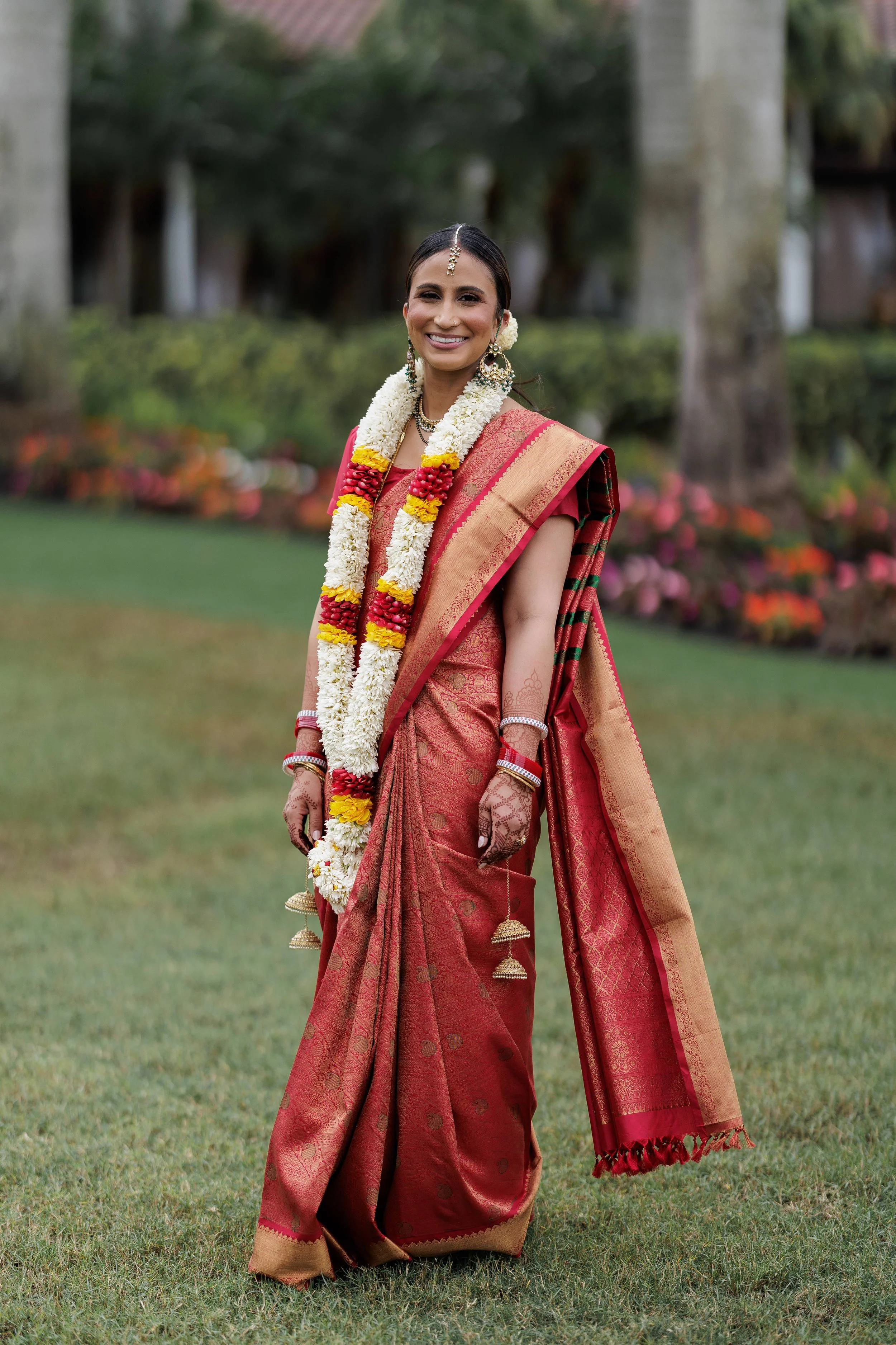 A woman in traditional Indian attire standing outdoors, smiling, wearing a red and gold saree, with floral jewelry and henna on her hands.