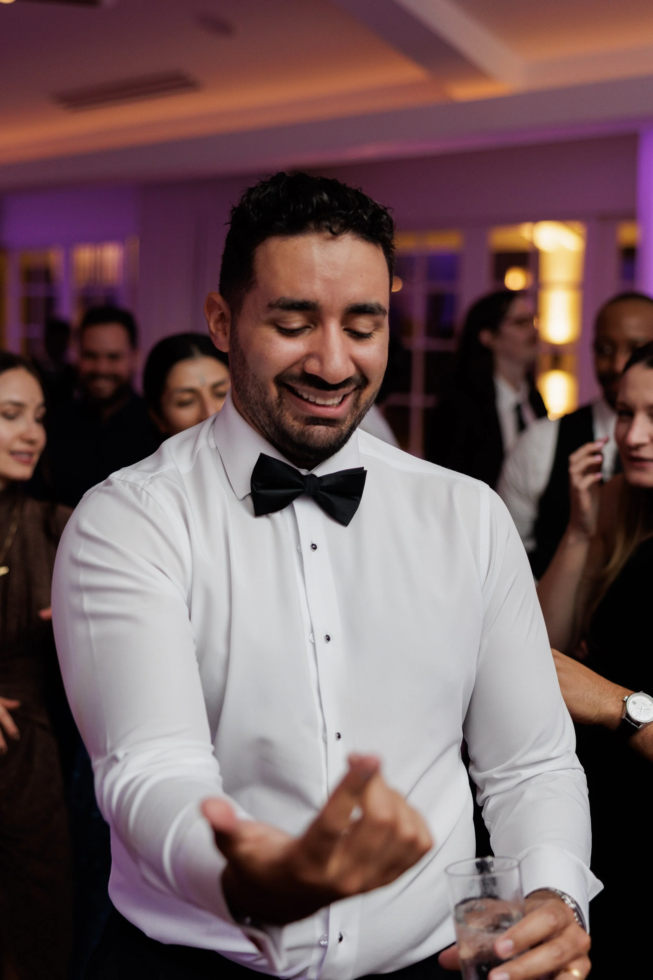 A man in a white dress shirt and black bow tie smiling at a party or celebration, holding a drink in his hand with people gathered around him, colorful lighting in the background.