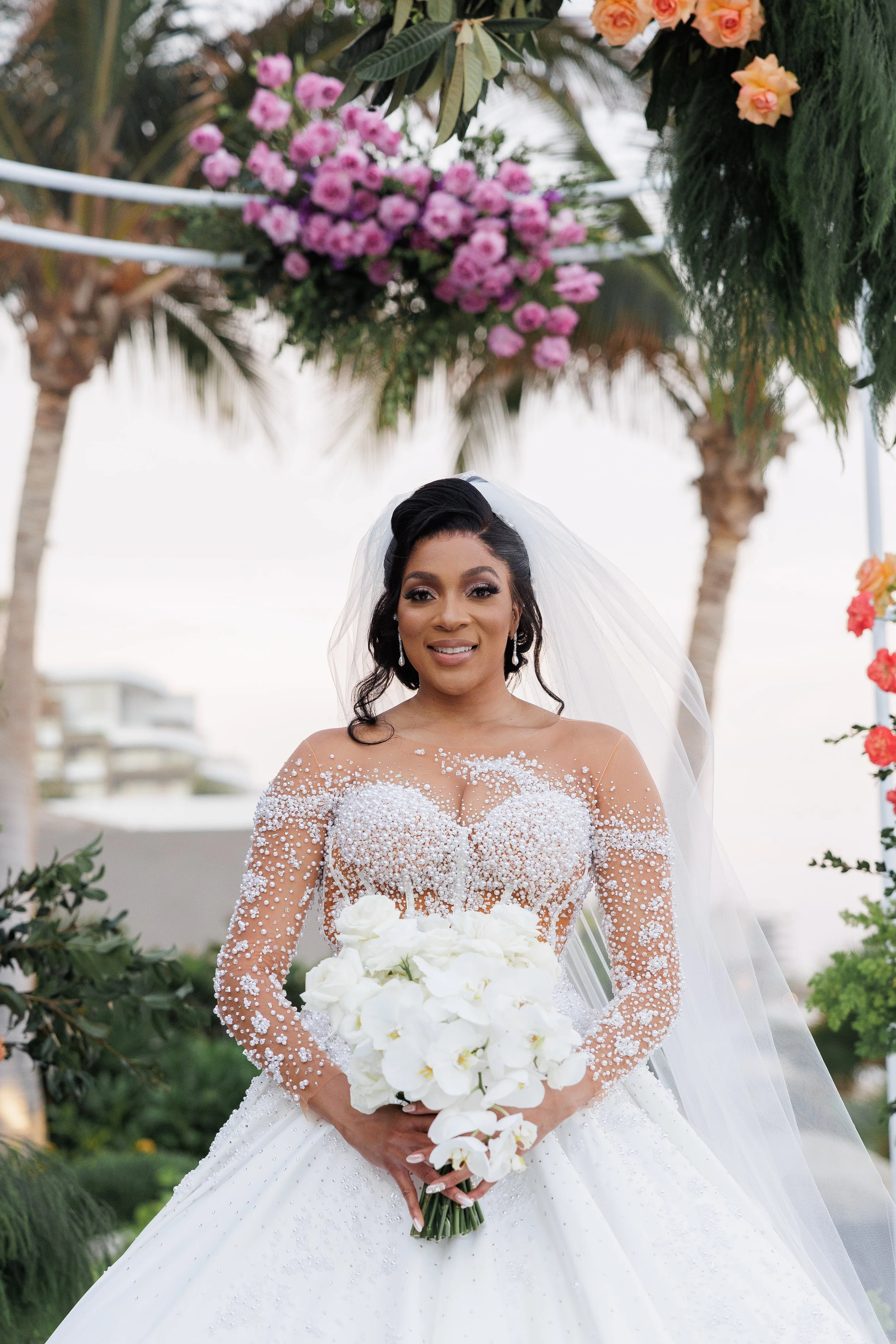 A bride in a wedding dress standing outdoors, holding a bouquet of white flowers, with palm trees and floral decorations in the background.