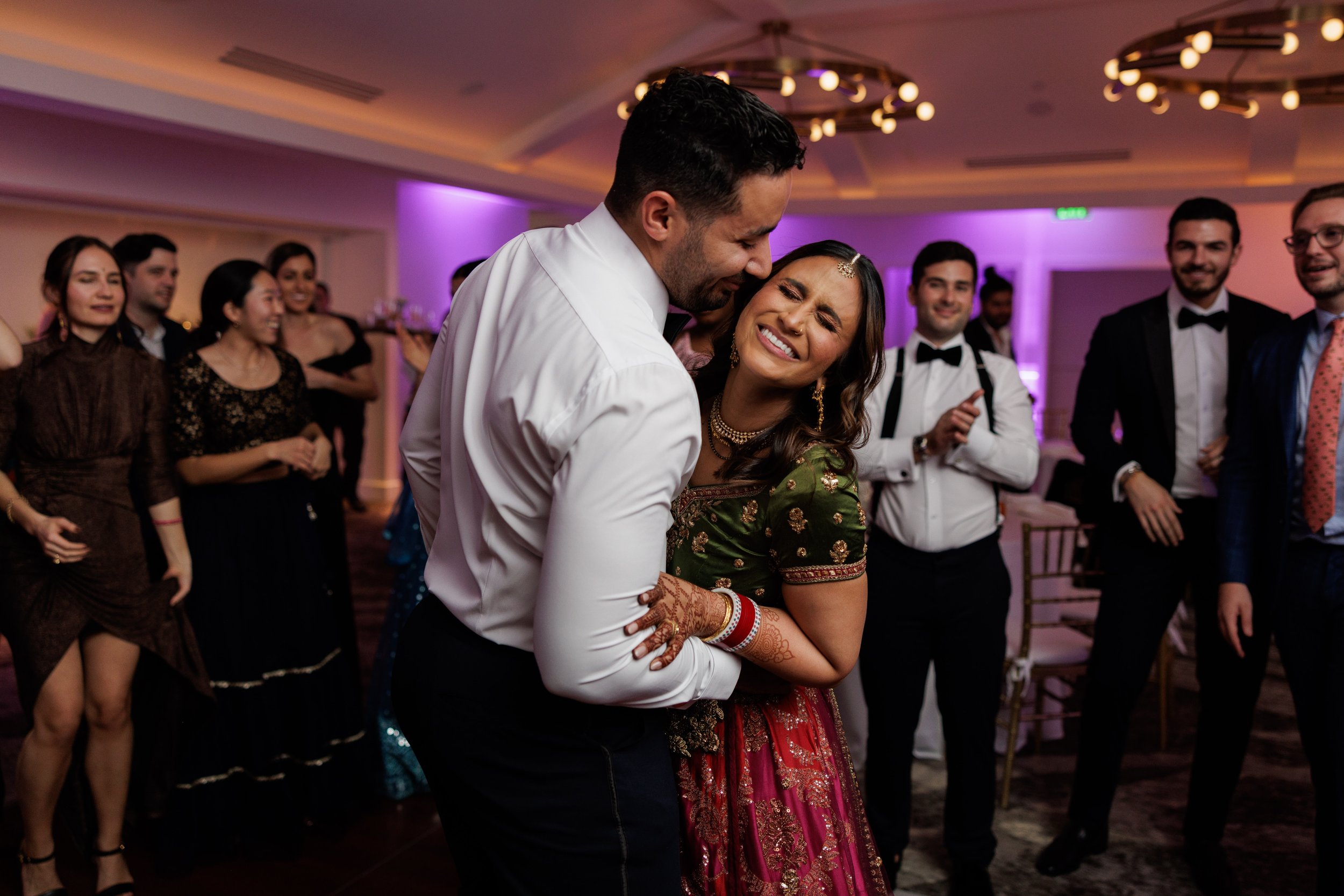 A happy couple dancing at a wedding reception, surrounded by smiling guests in formal attire, in a decorated indoor venue with purple lighting.