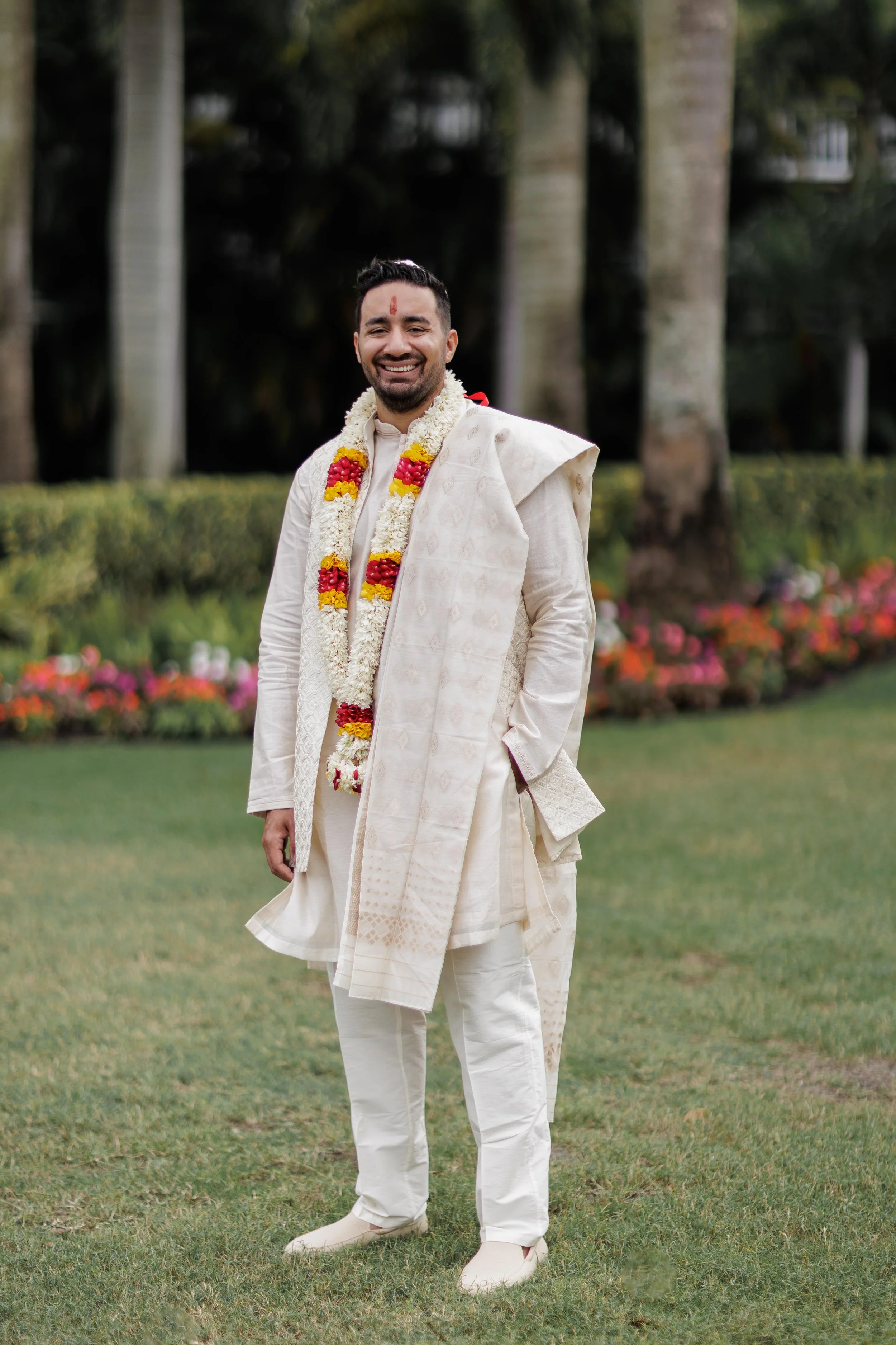 A smiling man dressed in traditional Indian attire, wearing a cream-colored kurta and dhoti, with a shawl draped over his shoulder. He has a garland of flowers around his neck and a red mark on his forehead, standing outdoors on grass with trees and 