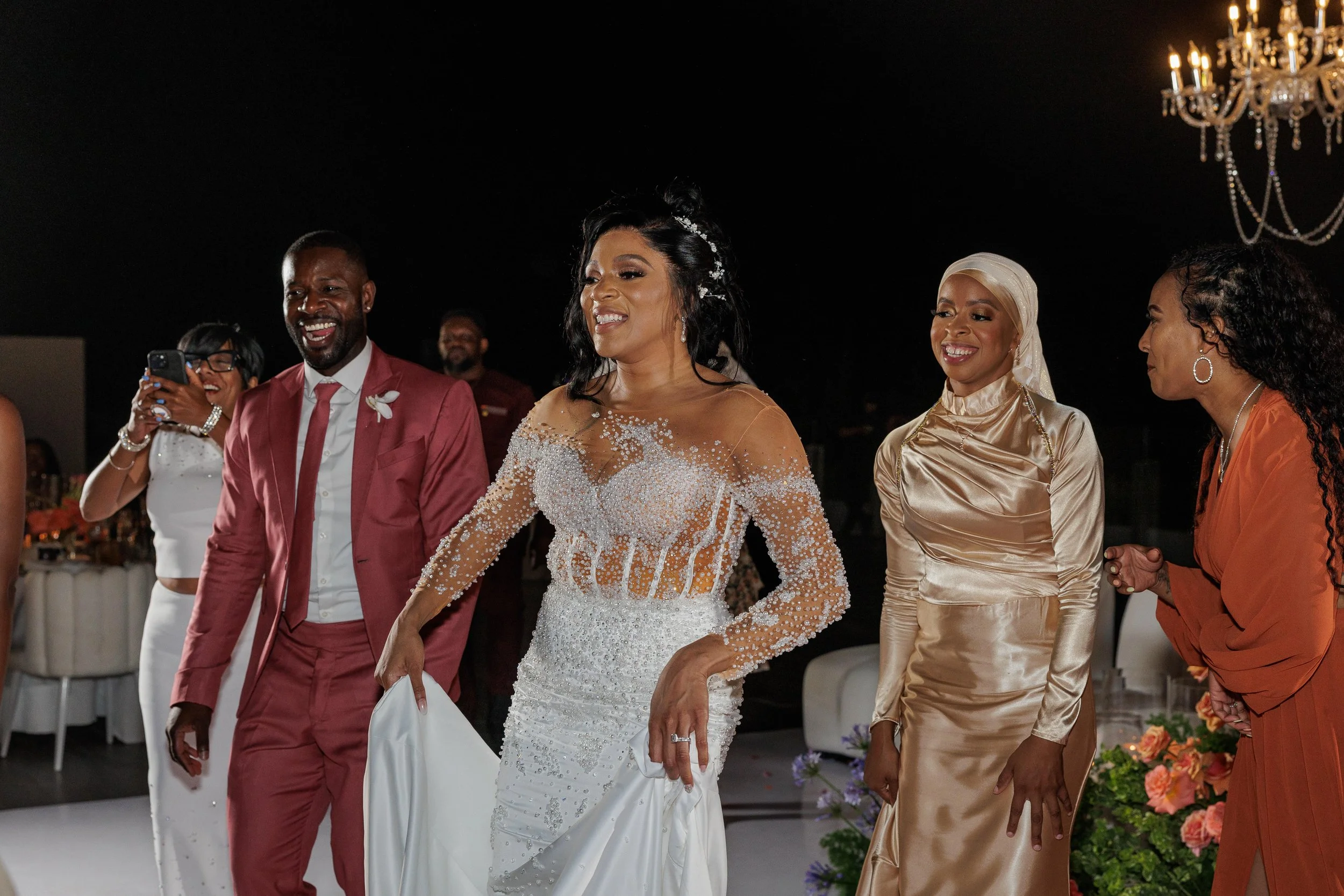 A bride in a white wedding dress with beadwork and a long train, smiling and holding her dress, at her wedding reception with guests around her.