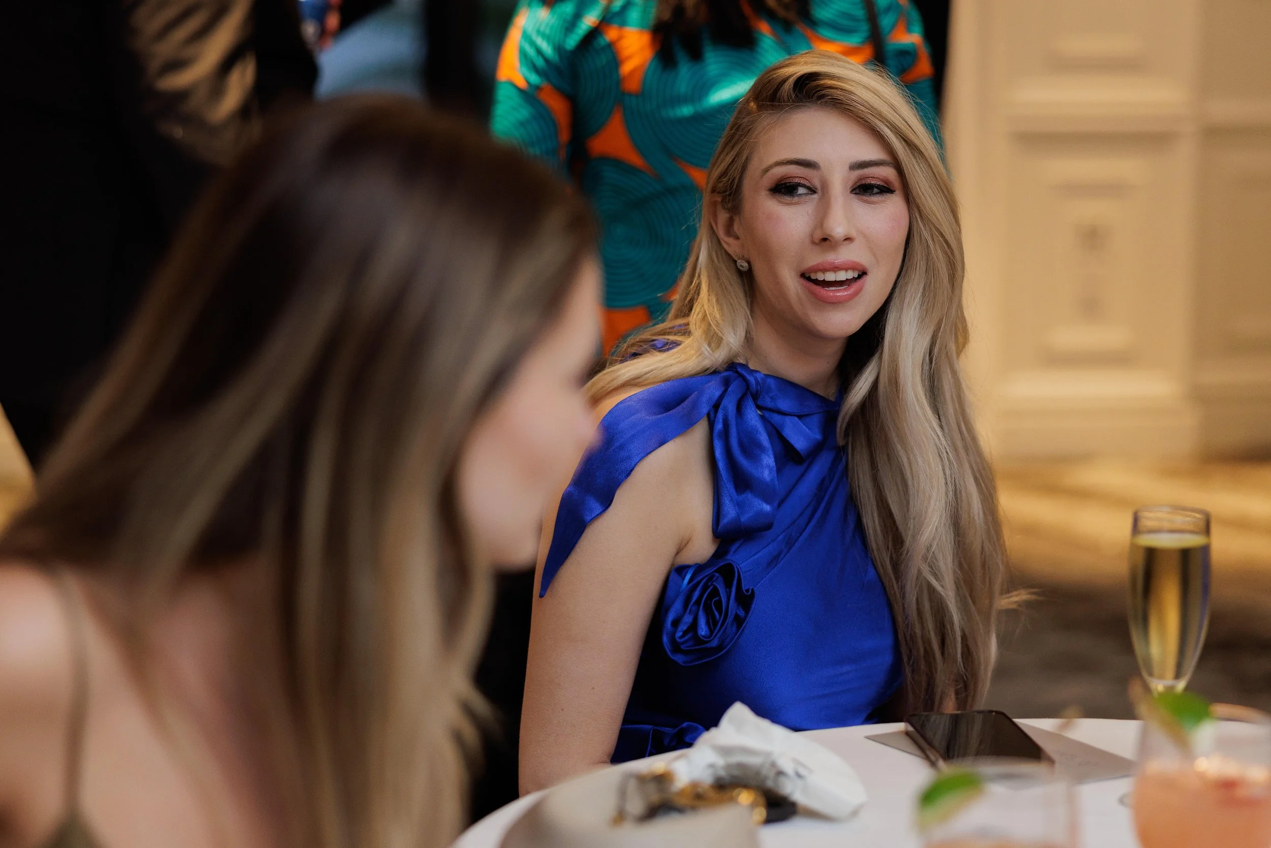 A woman with long blonde hair wearing a blue dress with bow and rose details, speaking at a formal event or dinner, with a glass of champagne and a smartphone on the table.