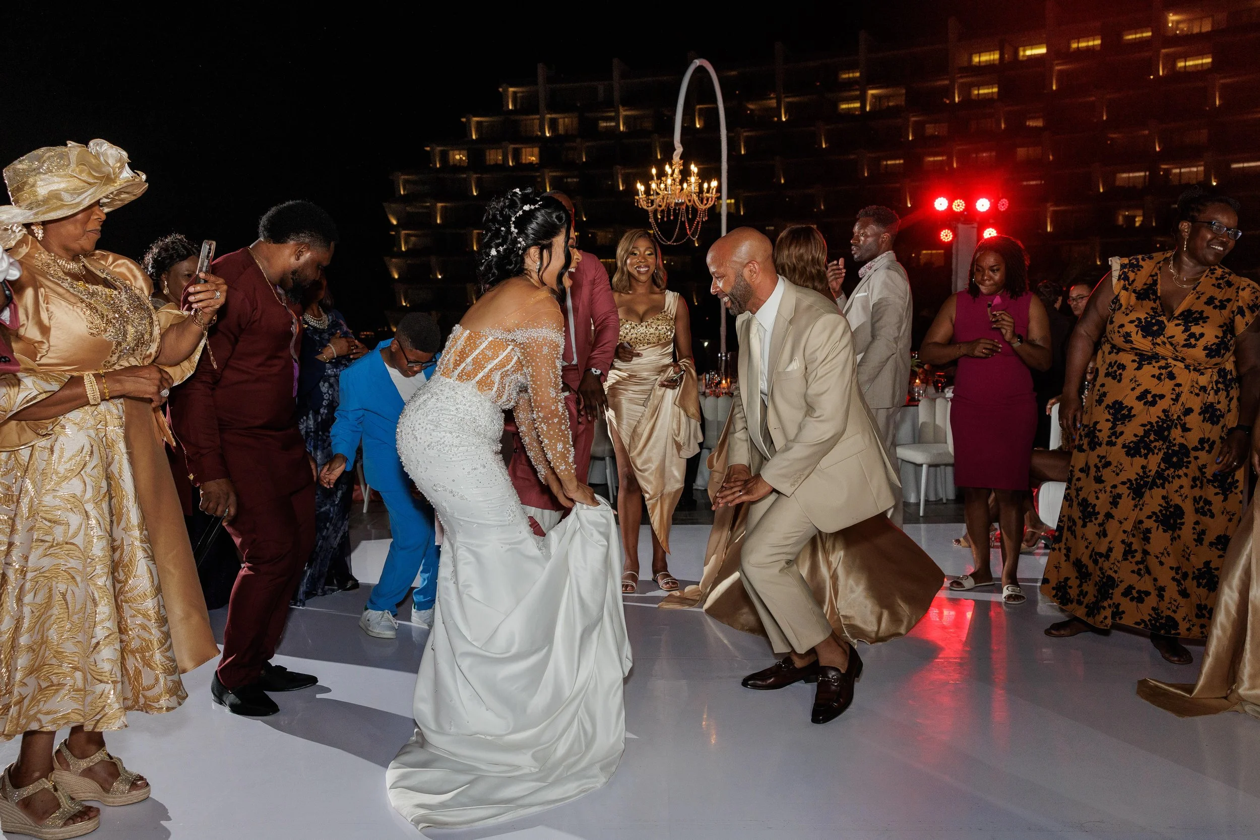People dancing at a wedding reception outdoors at night, with a large chandelier and city buildings in the background.