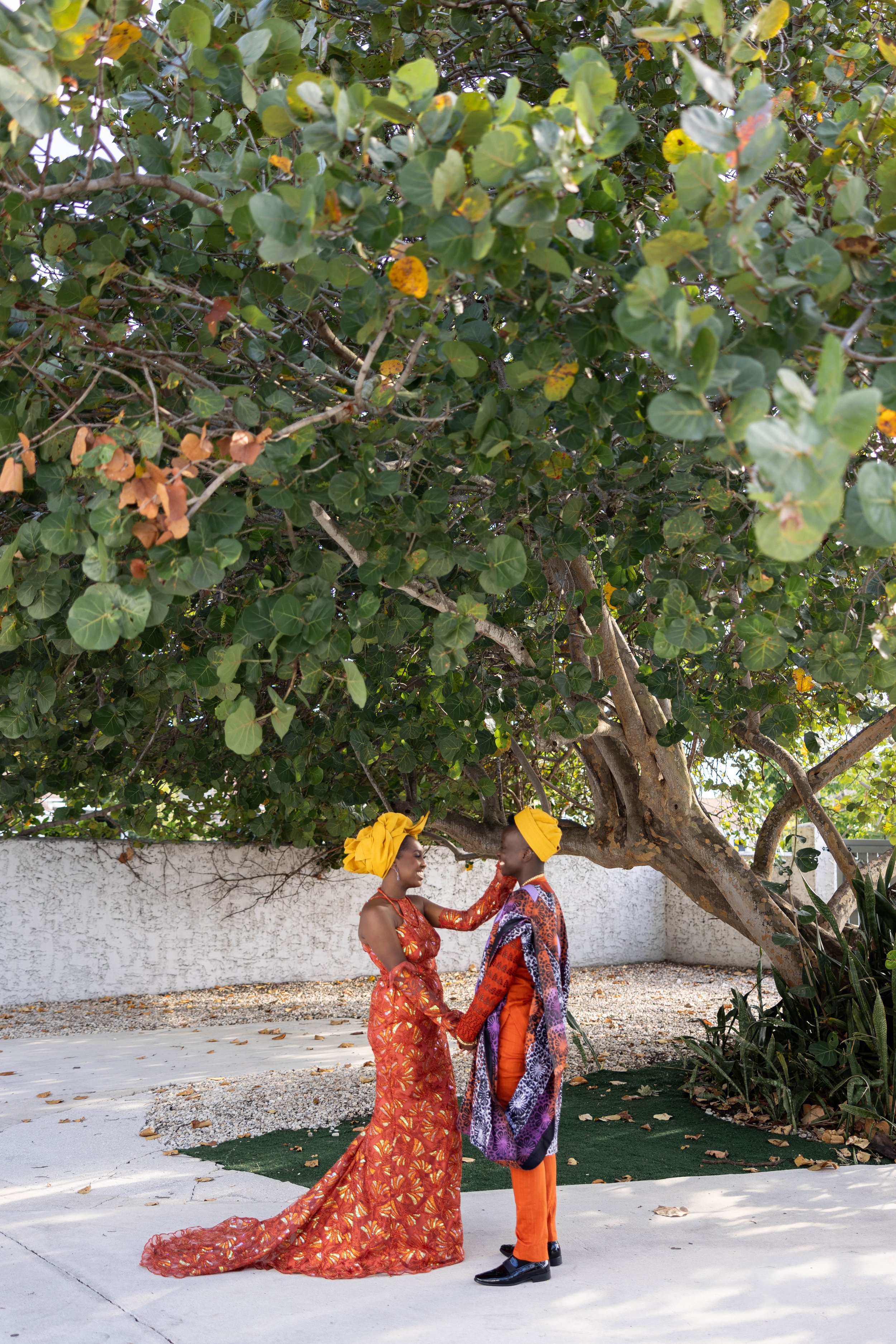 A man and woman dressed in traditional African attire, standing and holding hands under a large leafy tree, outside during the day.