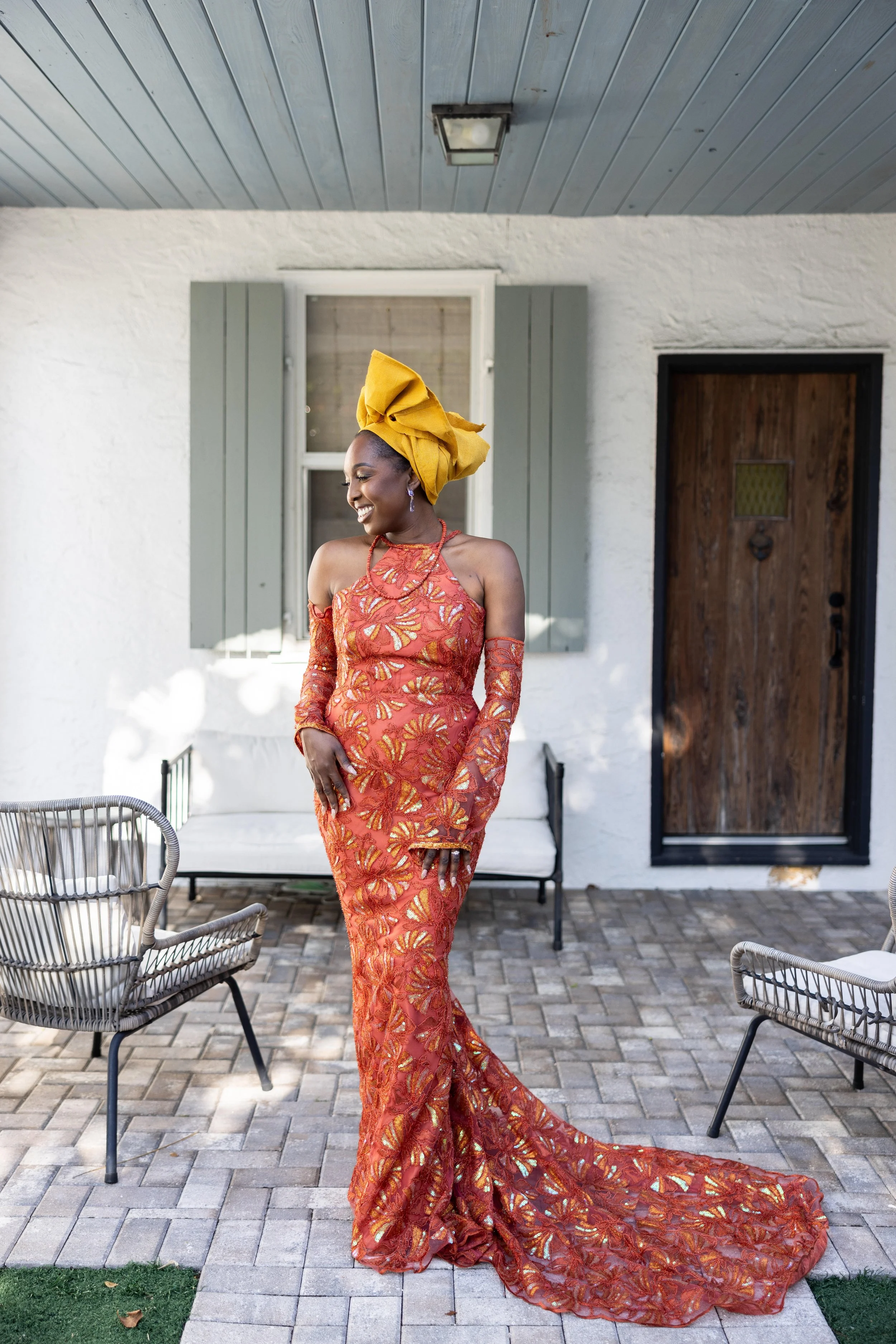 Woman wearing a vibrant orange and gold patterned dress, yellow headwrap, smiling, standing outdoors on a patio with furniture and a white house in the background.