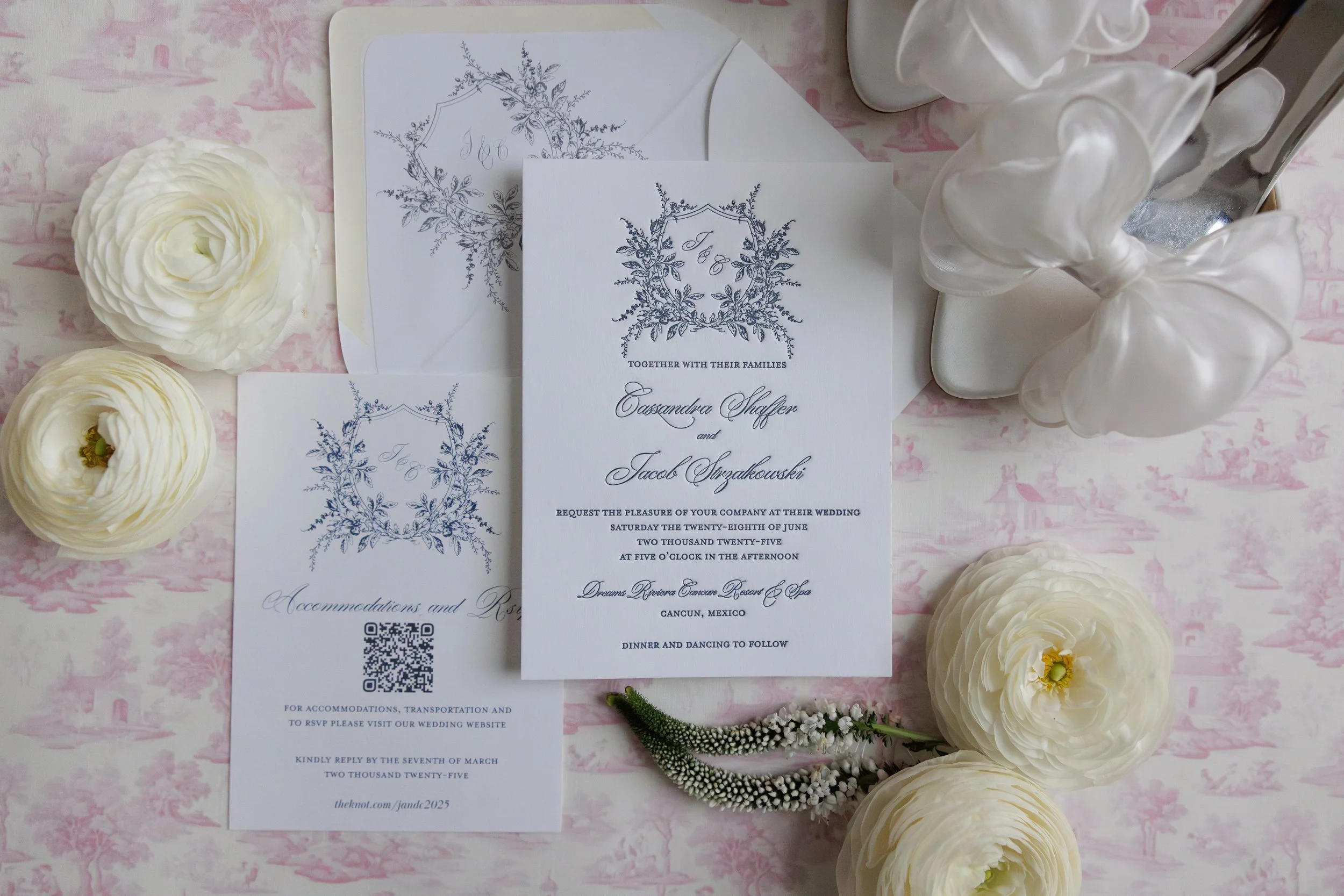 Wedding invitation suite with floral design, white flowers, and white balloon decorations on pink patterned tablecloth.