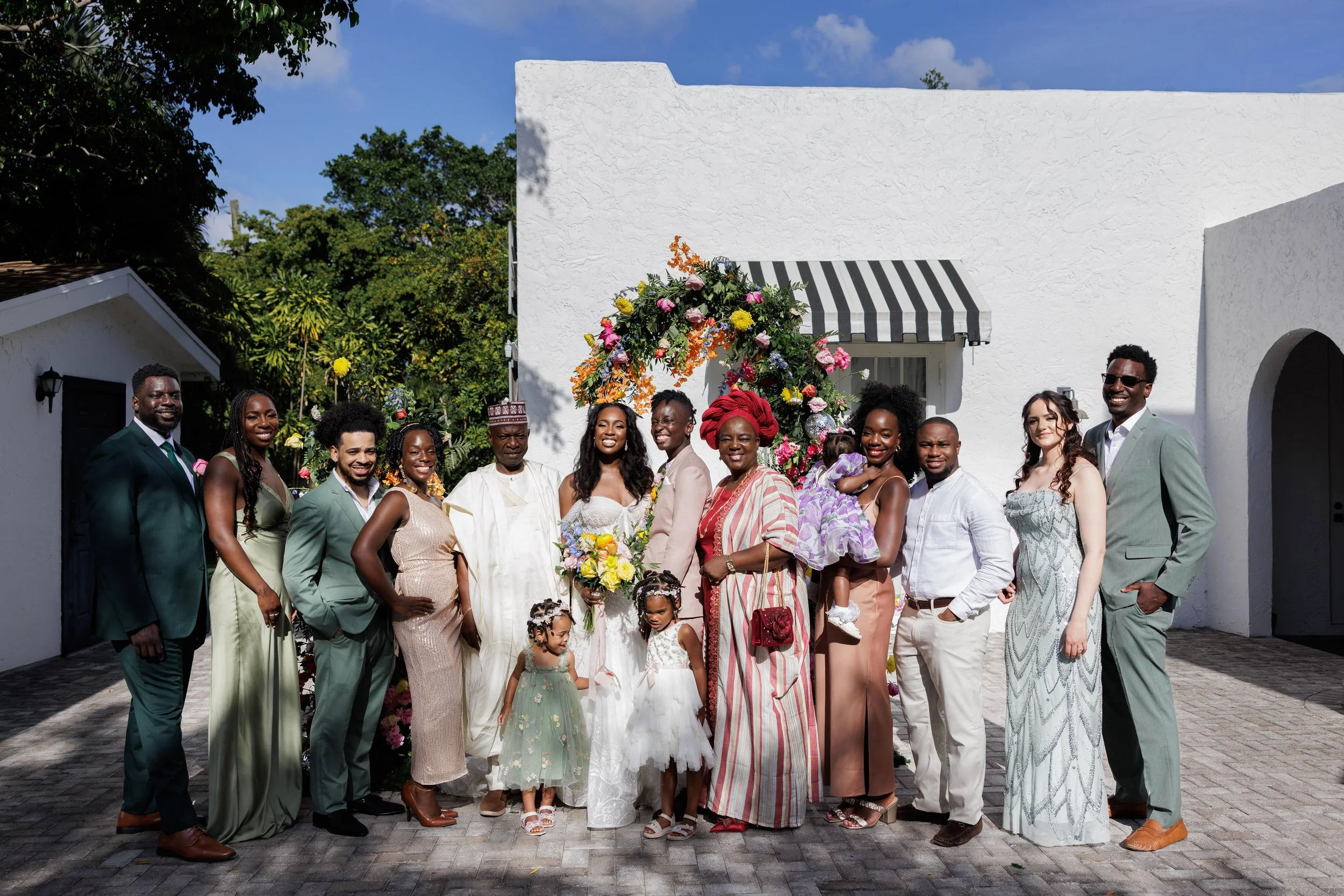 Group of people at a wedding celebration standing outside in front of a white building with a floral arch, sunlit sky, and greenery in the background. The group includes men, women, and children dressed in formal attire, smiling for the photo.