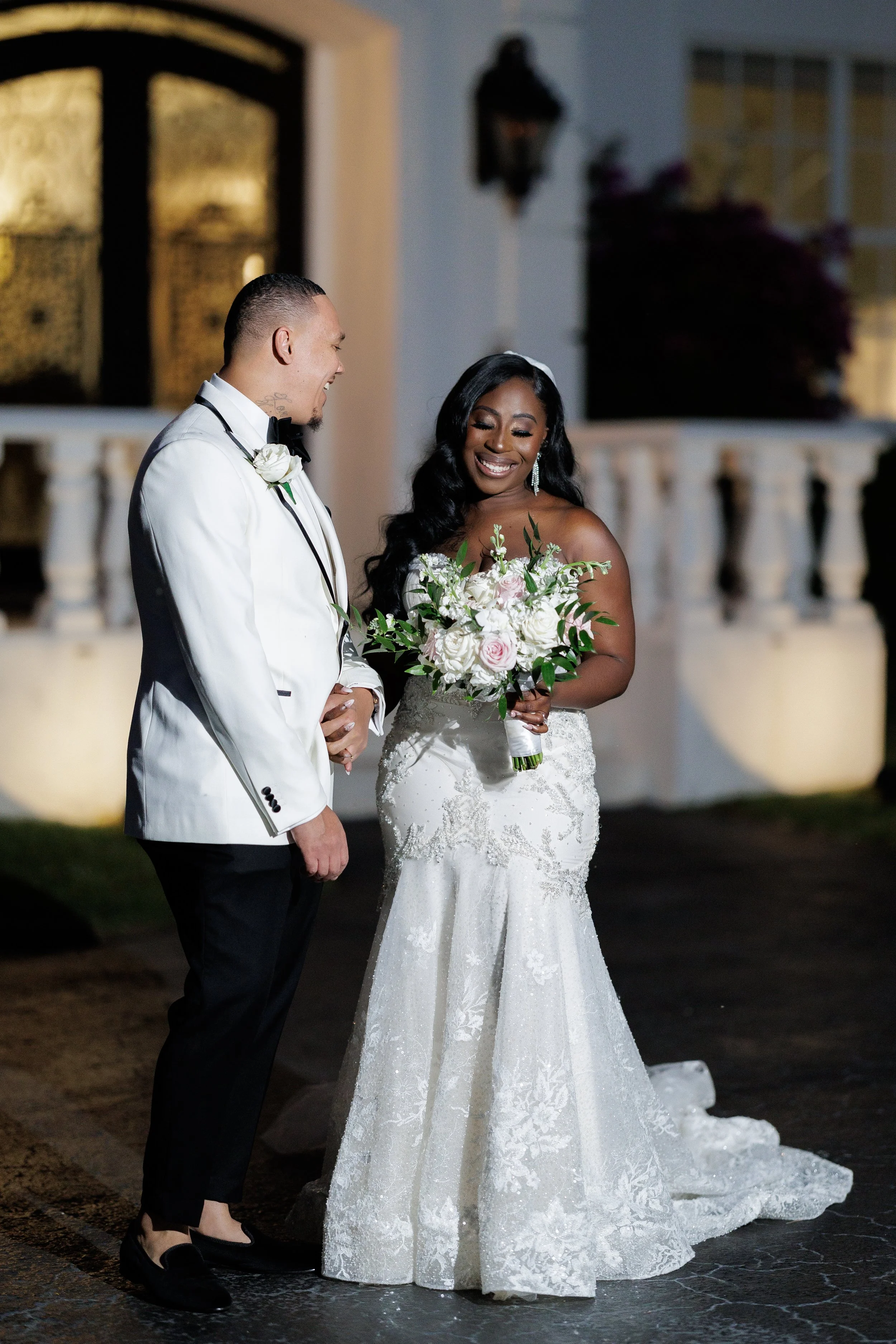 A bride and groom smiling at each other during their wedding reception outside at night. The bride wears a white wedding gown and holds a bouquet of white and pink flowers. The groom wears a white tuxedo jacket with black pants and shoes.
