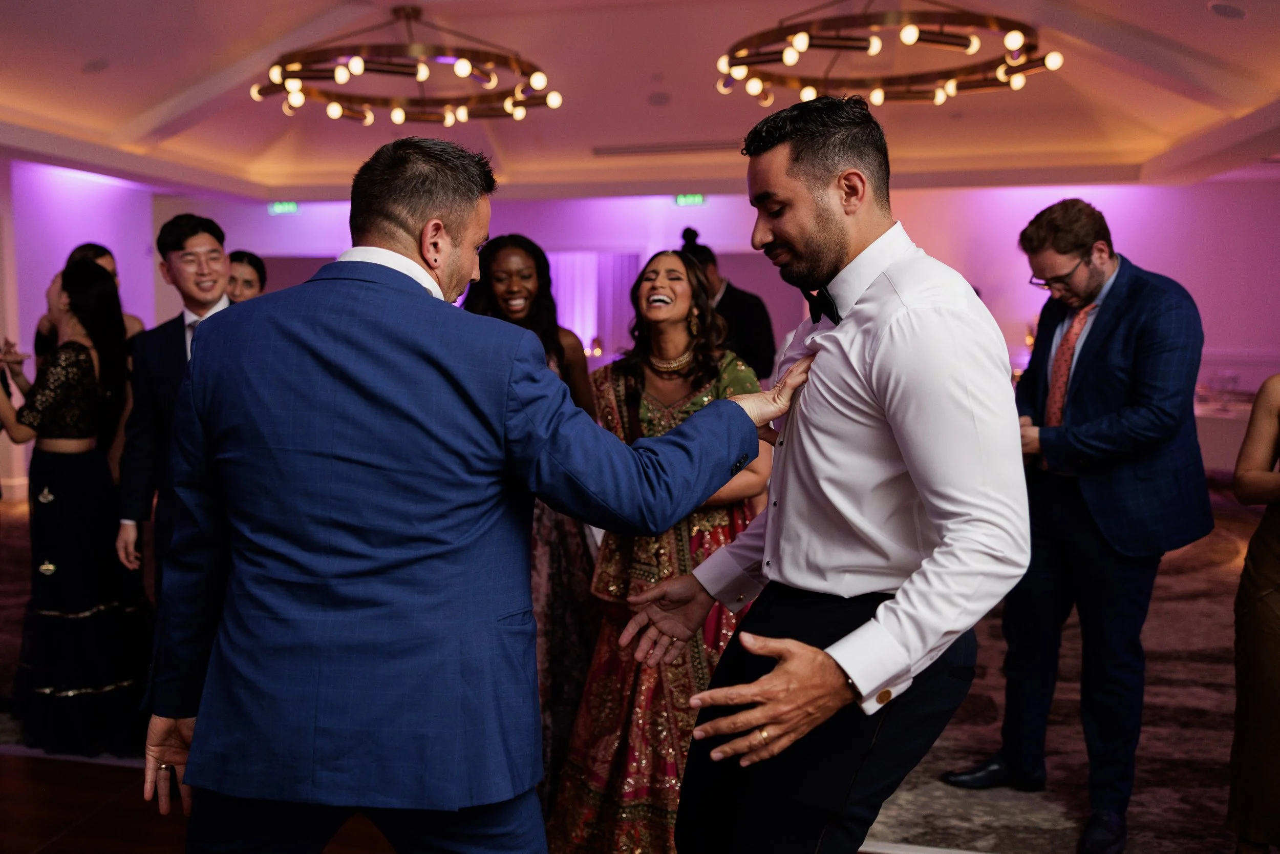 People dancing and enjoying themselves at a celebration, with the focus on two men dancing closely in the foreground, one in a blue suit and the other in a white shirt with a black bow tie, in a warmly lit event space.