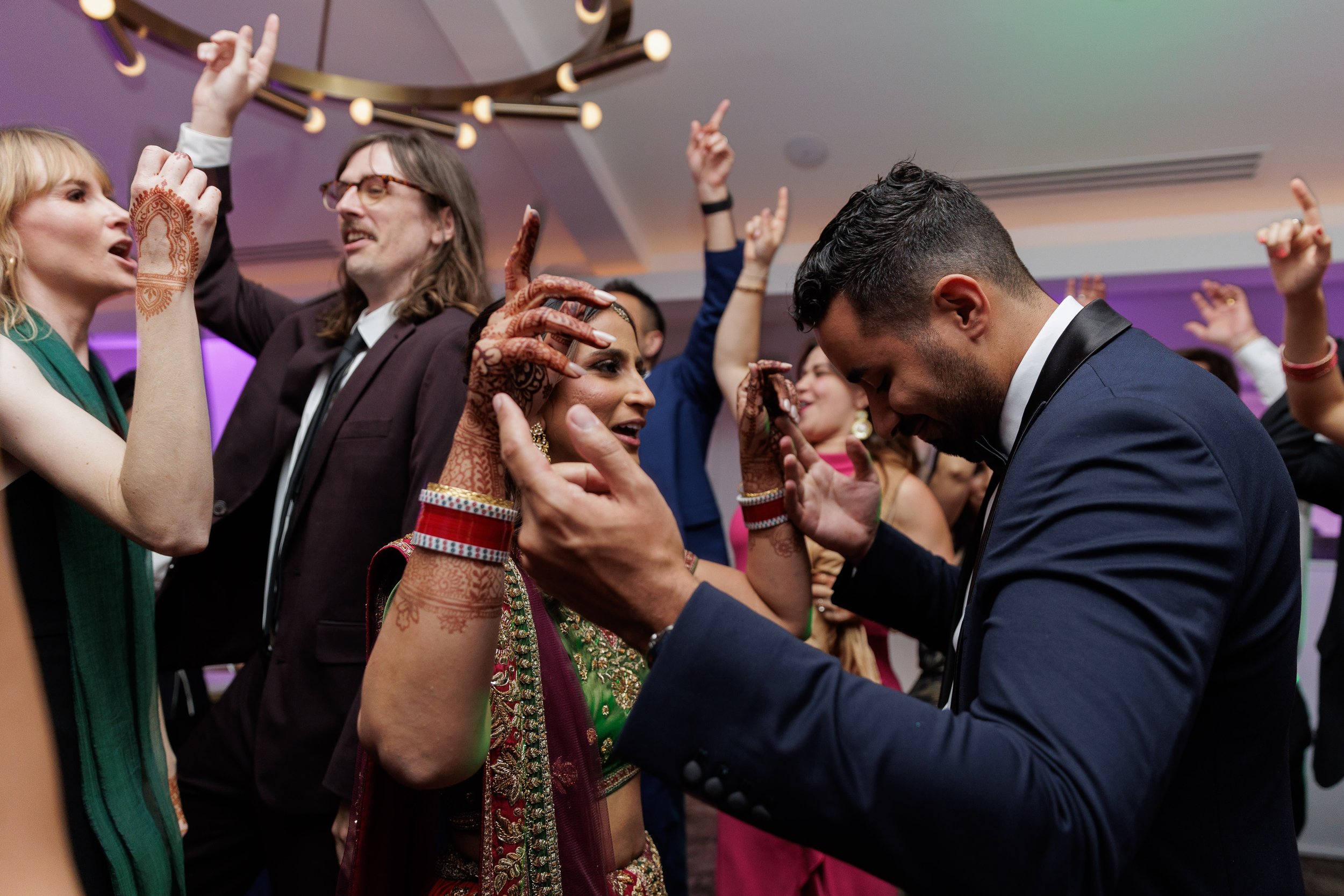 People dancing and celebrating at a wedding reception, with a bride in traditional Indian attire and groom in a tuxedo.