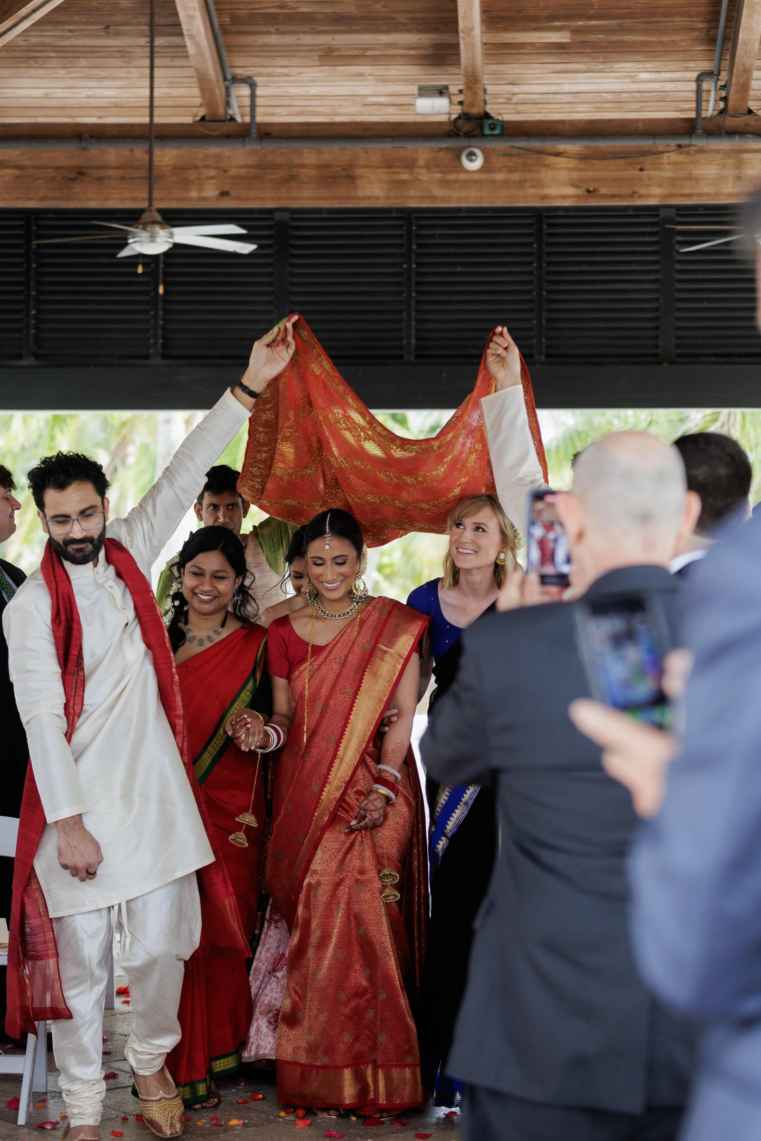 Indian wedding ceremony with women in traditional sarees and jewelry, and men in traditional attire, celebrating with a cloth held above the bride's head, while another person takes a photo.