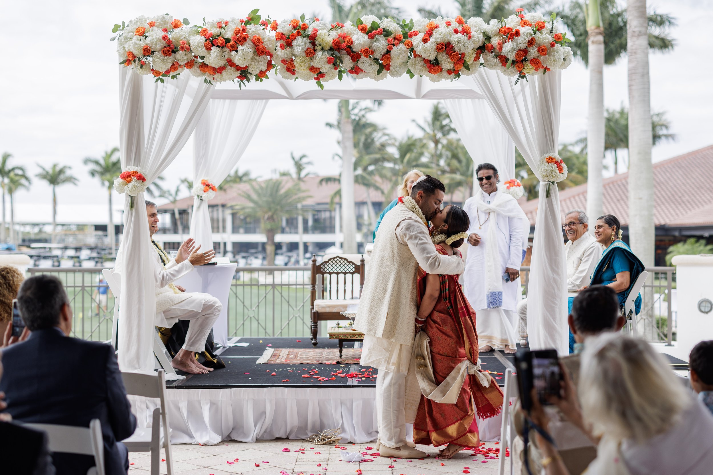 Indian couple getting married in a wedding ceremony outdoors under a floral canopy with white drapes, surrounded by family and friends, with a marina and palm trees in the background.