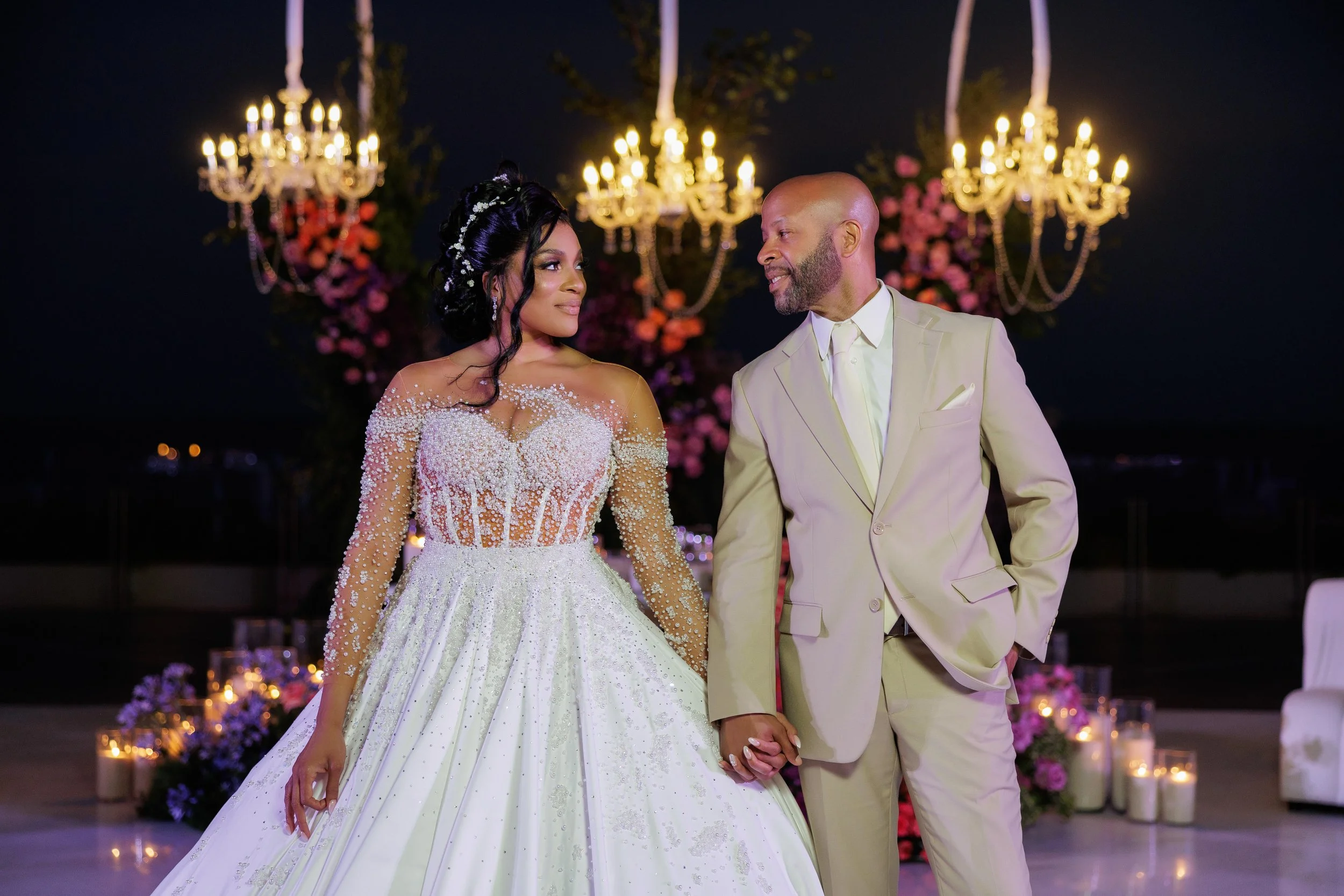 A bride and groom holding hands and looking at each other during their wedding reception, with chandeliers and flowers in the background.