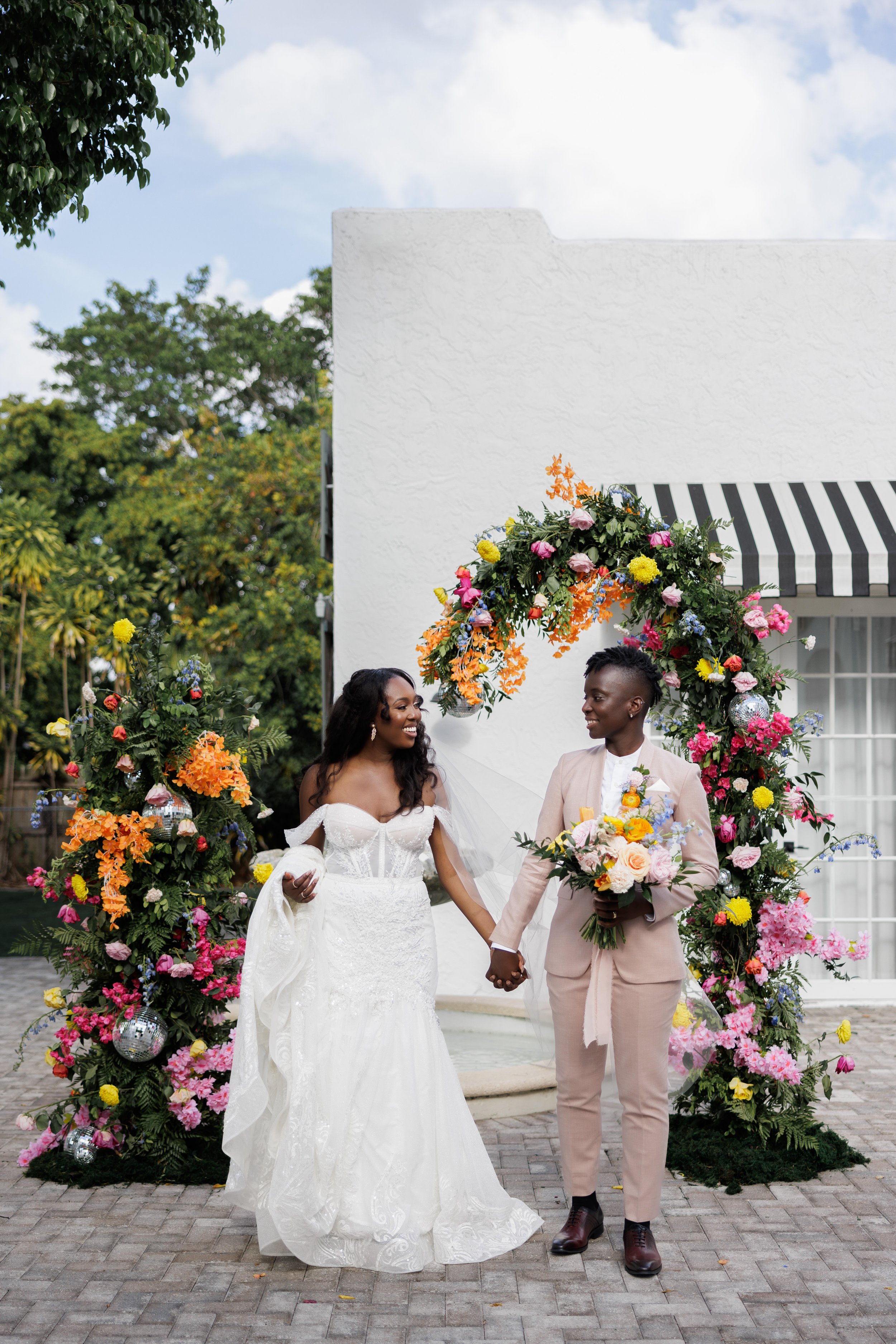 A same-sex couple holding hands and smiling at each other during their wedding ceremony, standing in front of a floral arch decorated with colorful flowers and disco balls, outdoors on a paved area with a white building and greenery in the background