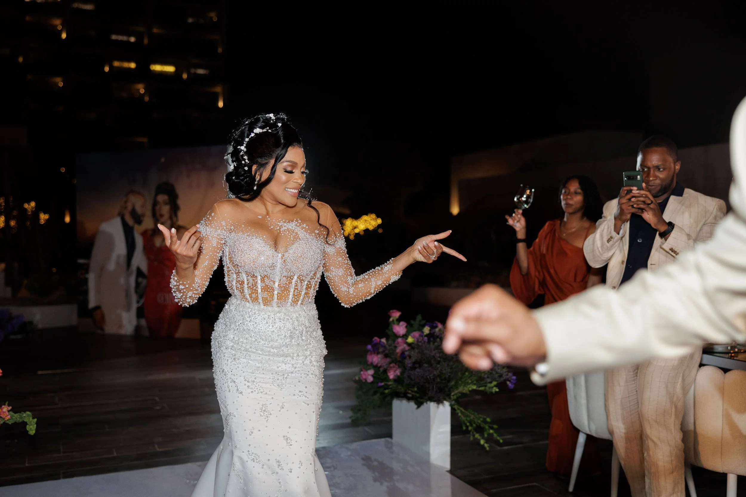 A bride in a decorated white wedding gown dancing at her wedding reception, with guests taking photos and videos of her.