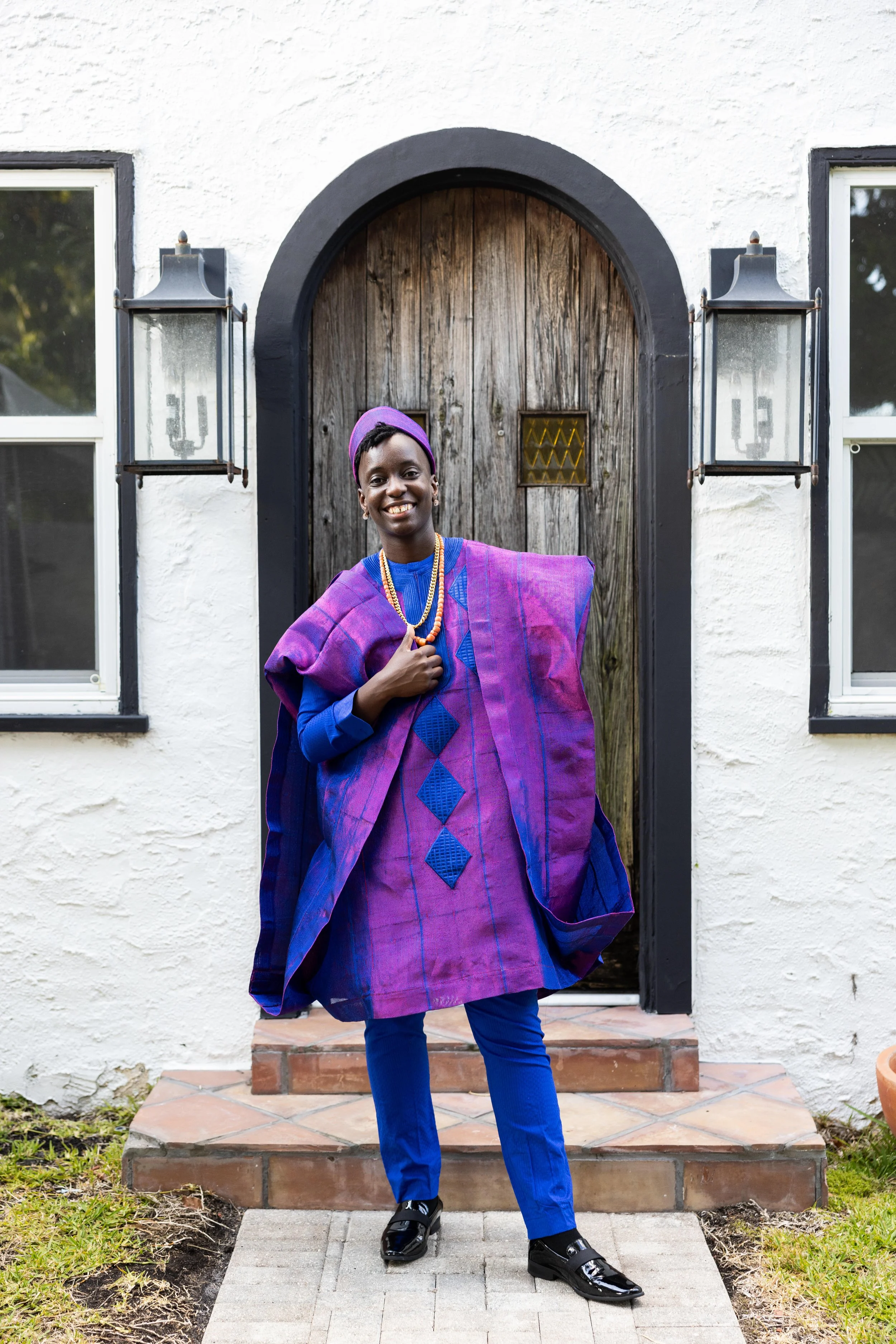 A woman wearing traditional African attire in blue and purple standing on a stone pathway in front of a wooden door painted black and white walls, flanked by two outdoor lanterns.