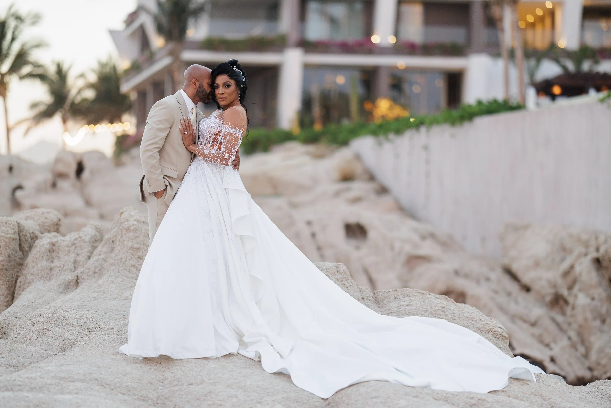 A bride and groom in wedding attire sharing an intimate moment on a rocky beach with a building in the background.