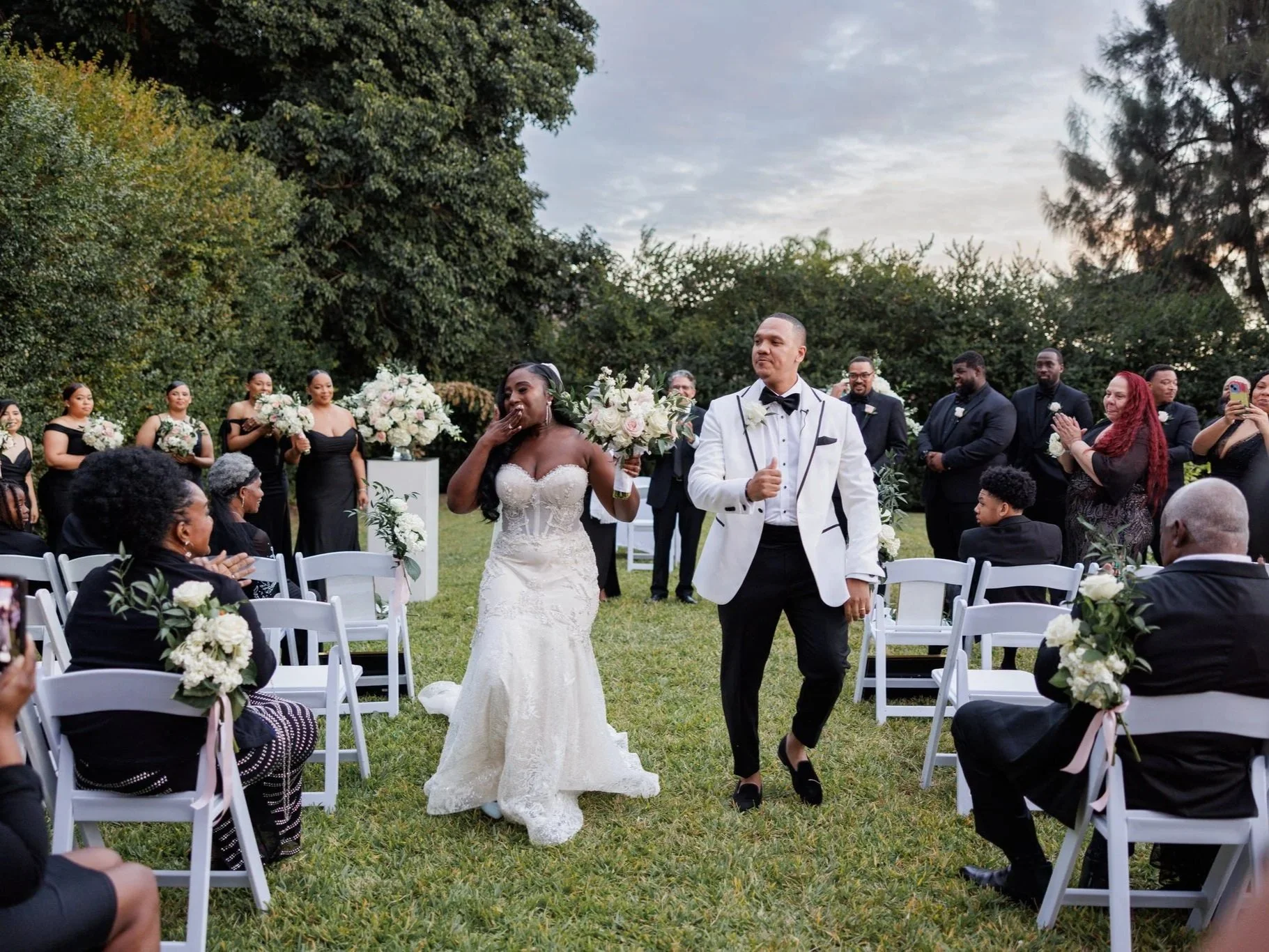 A bride and groom holding hands during their wedding, with the groom kissing the bride's forehead and the bride smiling, surrounded by tropical plants.