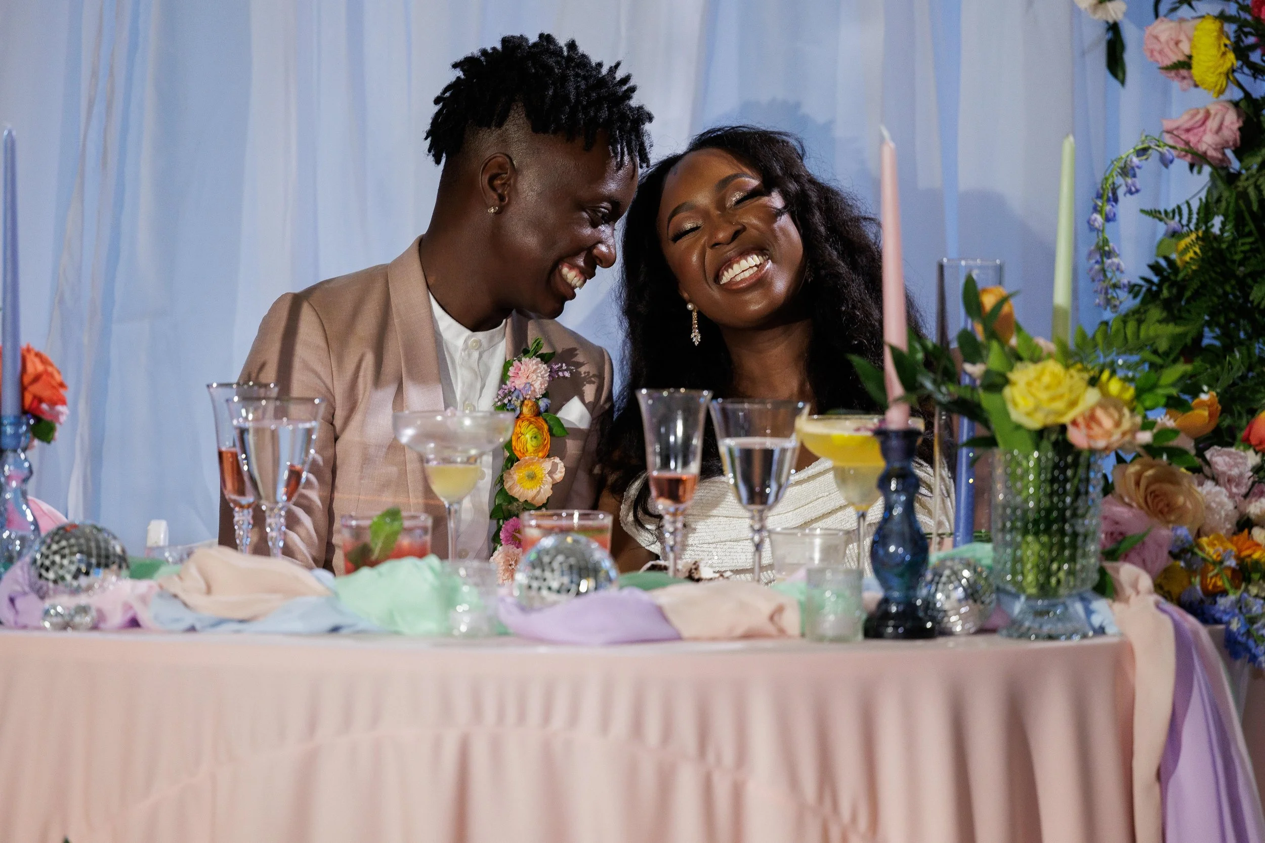 A smiling couple at a decorated table, possibly at a wedding reception, with colorful flowers and candles around them.