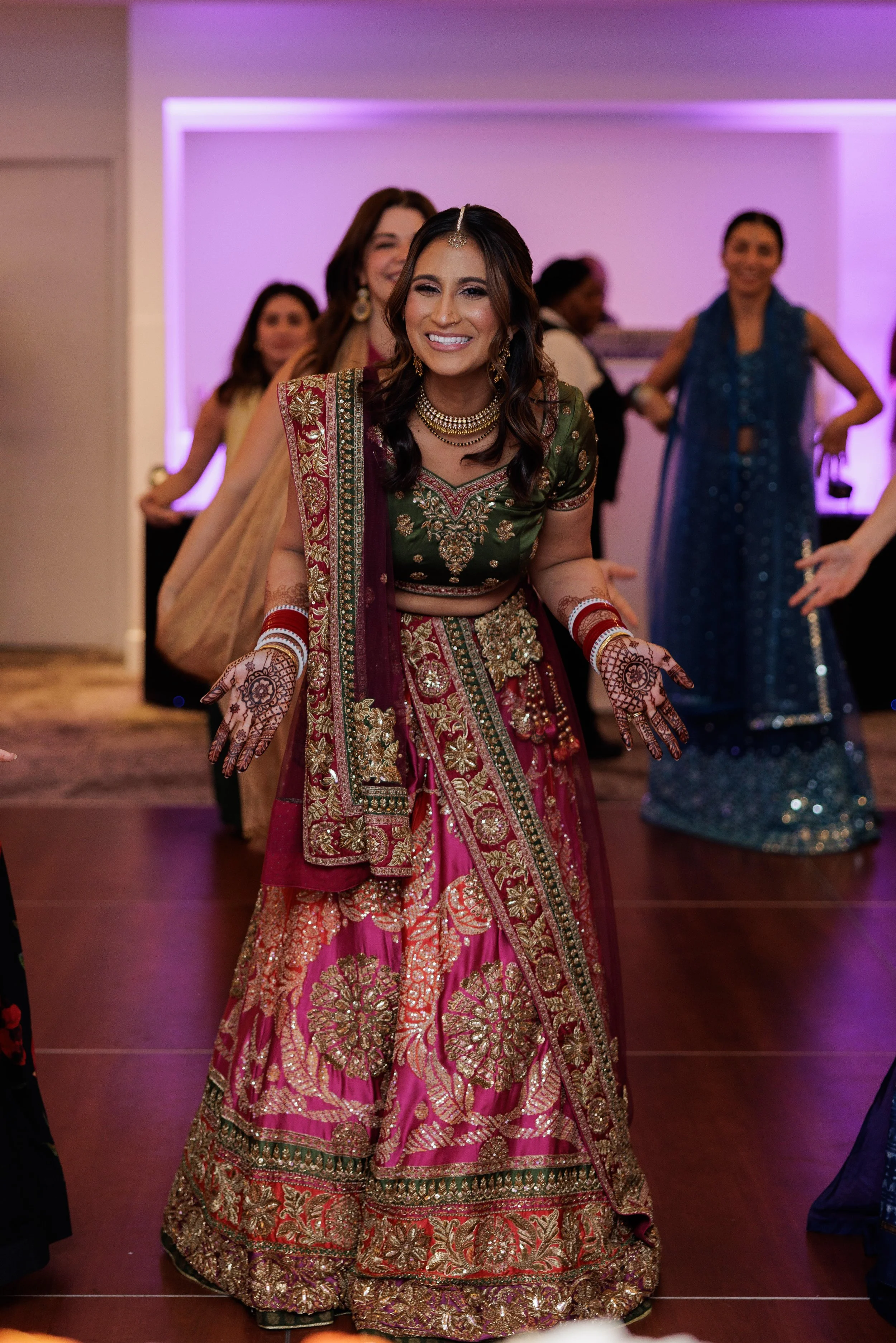 Woman dressed in traditional Indian attire at a festive event, surrounded by other women in colorful dresses, with purple lighting background.