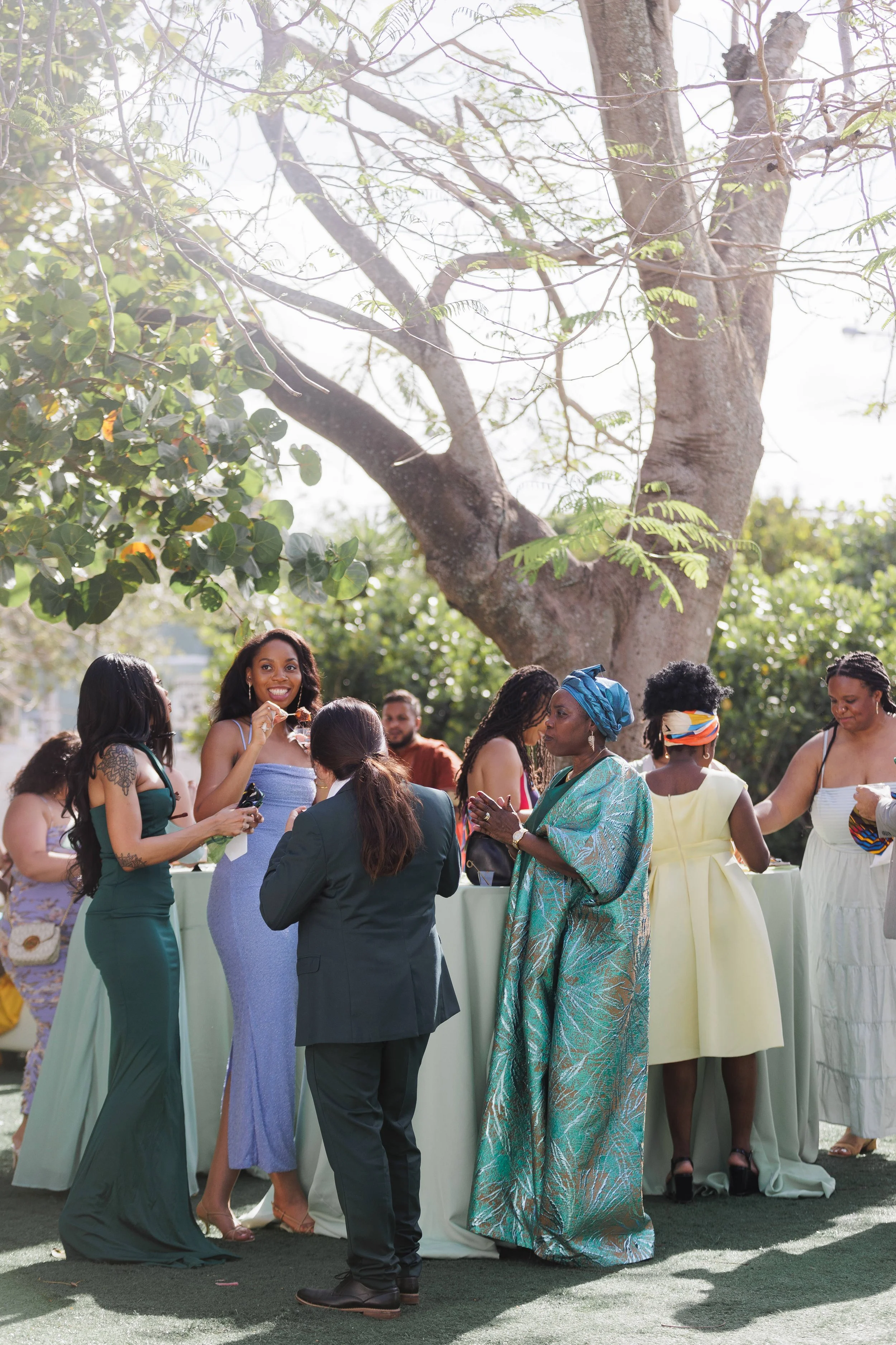 A group of women at an outdoor social gathering or celebration, standing around a tall table, under a large tree, enjoying conversation and drinks on a sunny day.