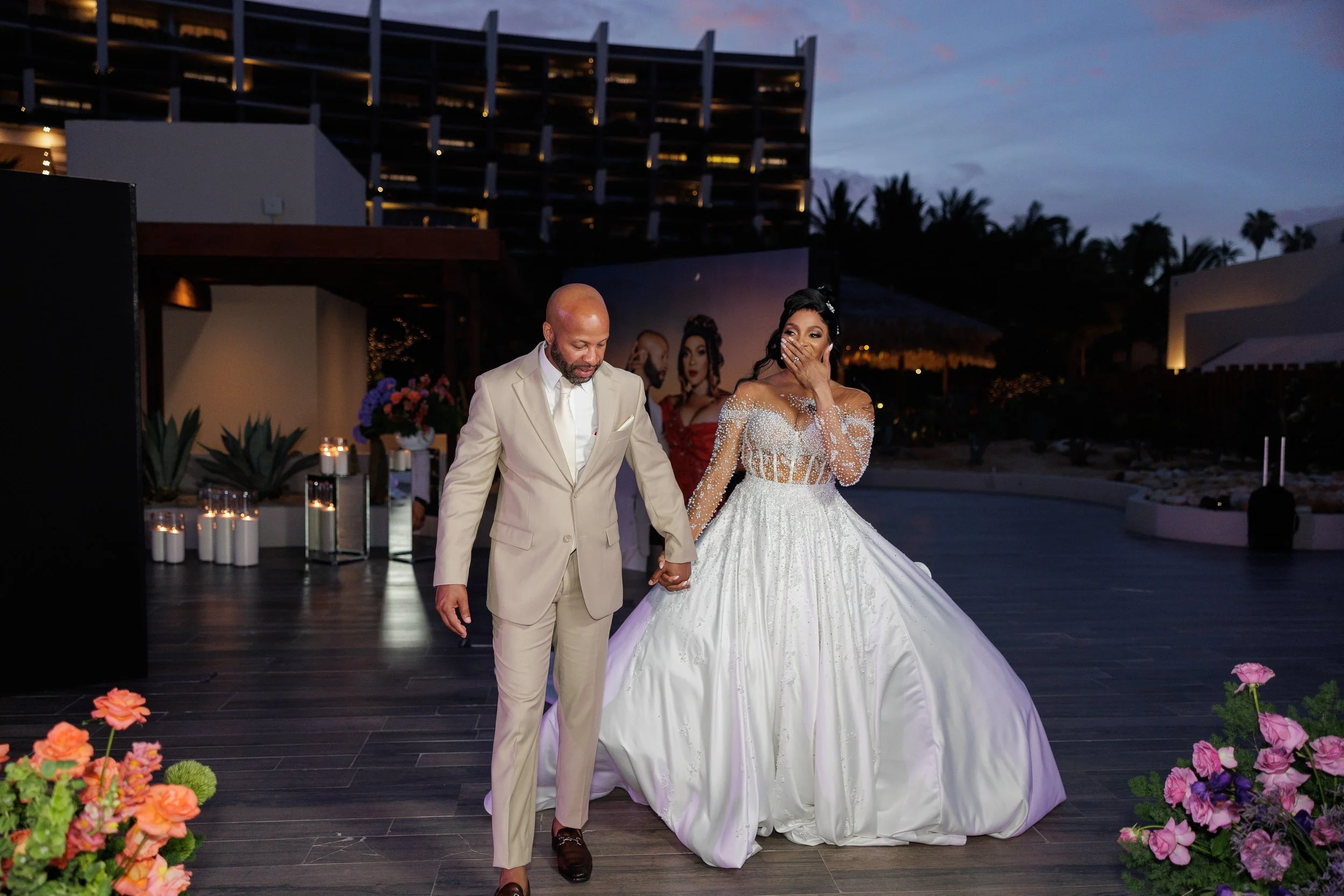 A bride in a sparkling white wedding gown and a groom in a beige suit stand hand-in-hand during their wedding reception outdoors at dusk, surrounded by floral arrangements and candles.