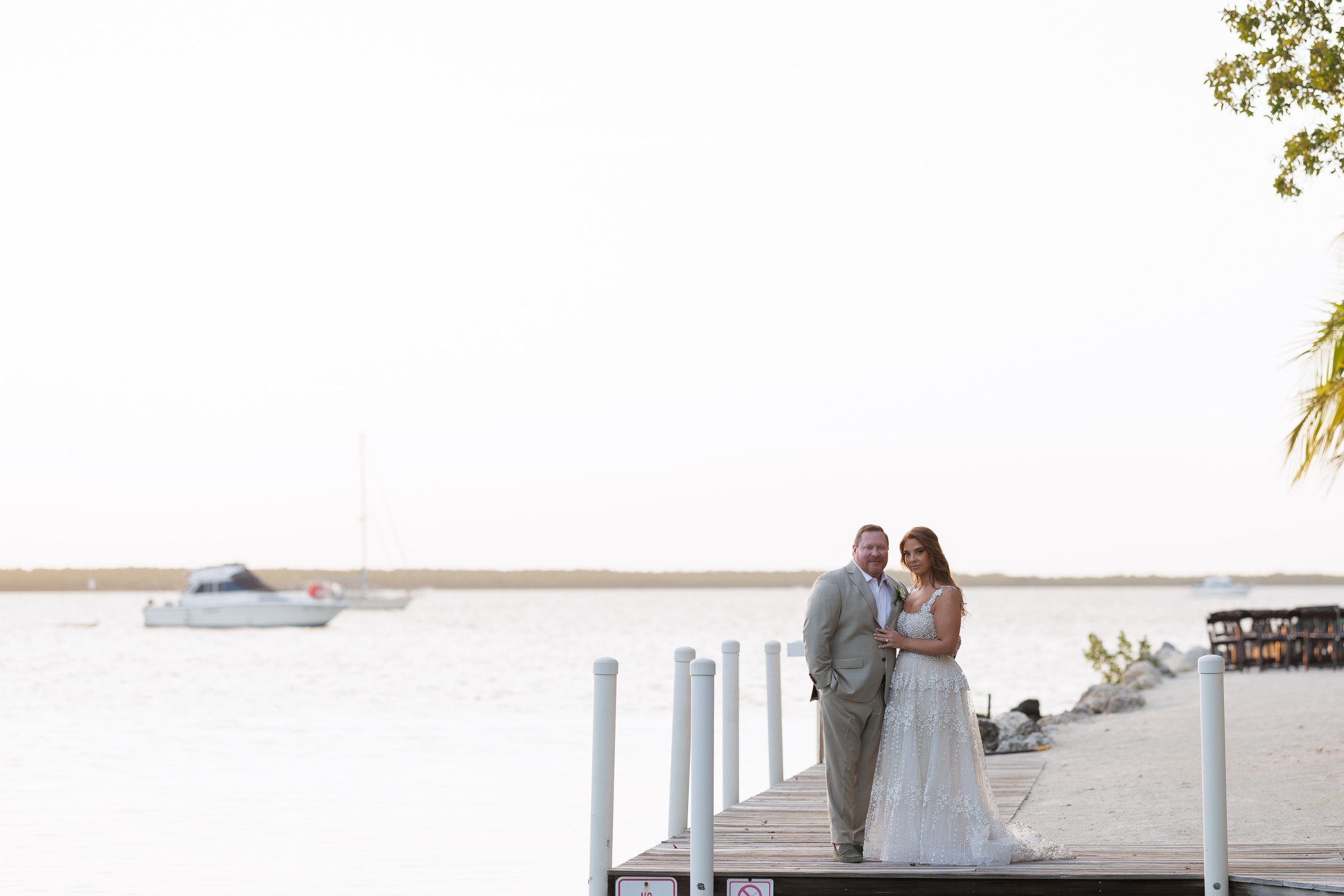 A bride and groom holding hands during their wedding, with the groom kissing the bride's forehead and the bride smiling, surrounded by tropical plants.