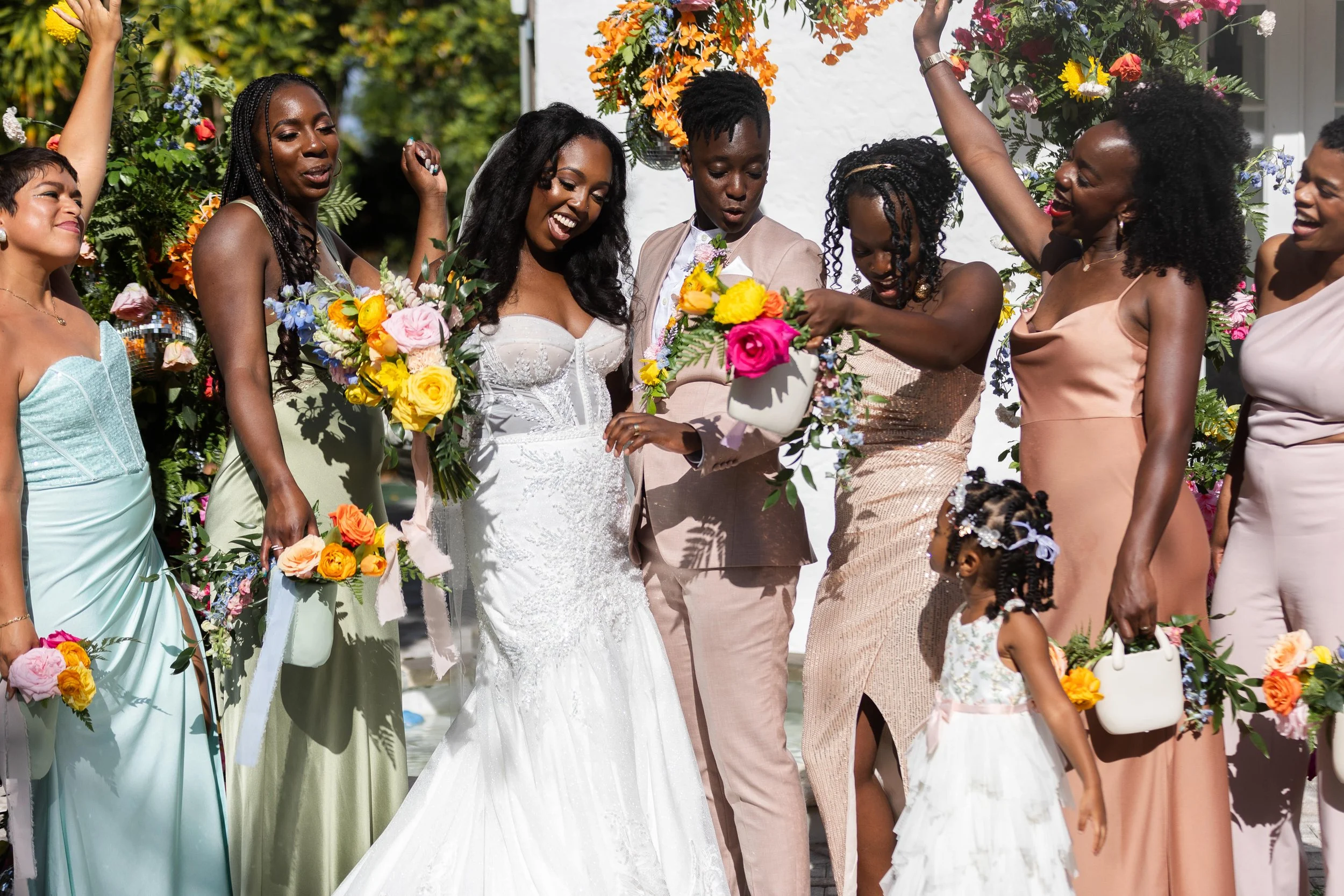 A group of women and a young girl celebrating at a wedding, holding bouquets of colorful flowers, with a bride in a white wedding dress at the center, outdoors with greenery and floral decorations.