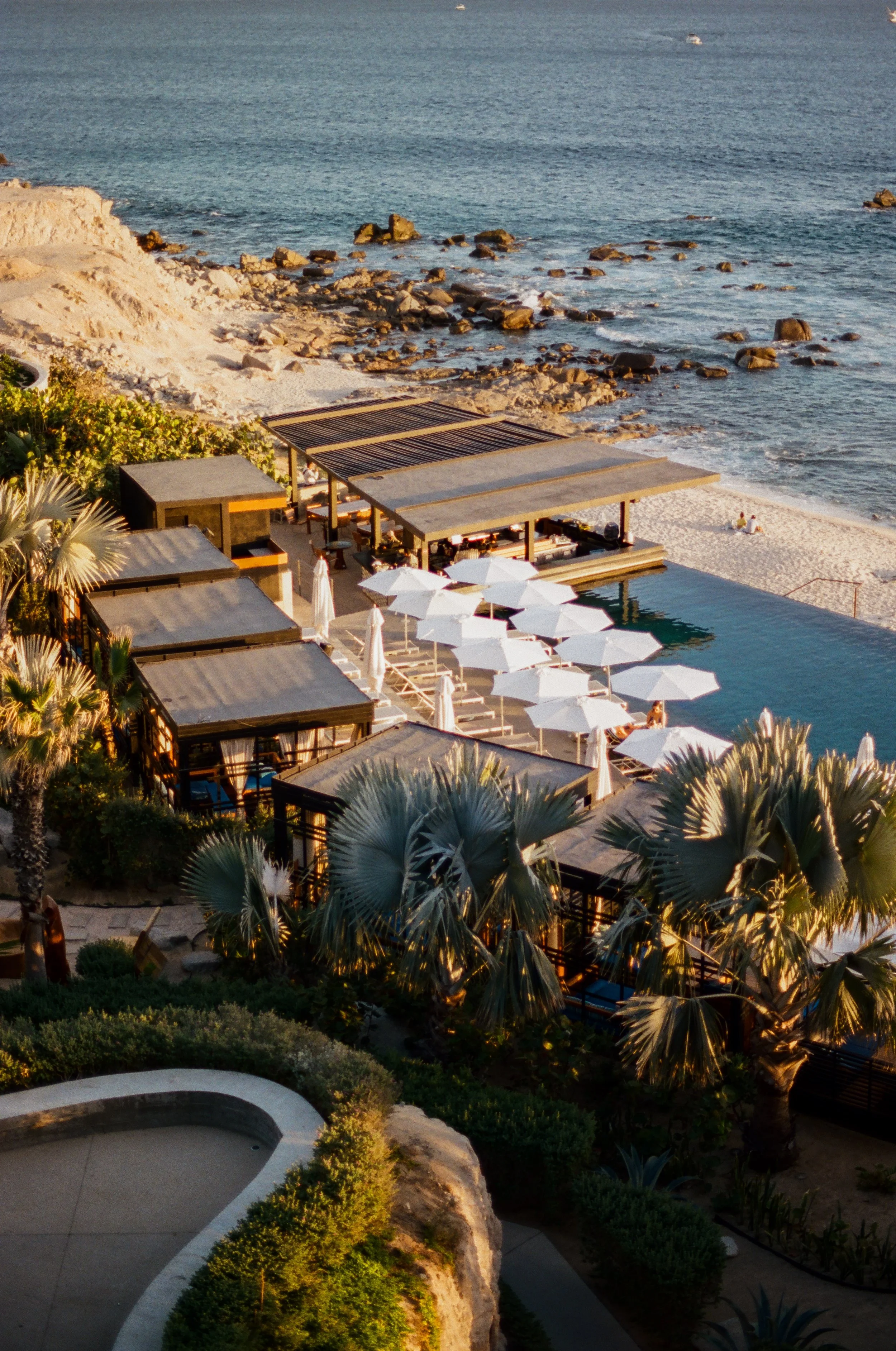 A seaside resort with cabanas, umbrellas, and lounge chairs by a pool, overlooking the beach and ocean with rocks and calm waves.