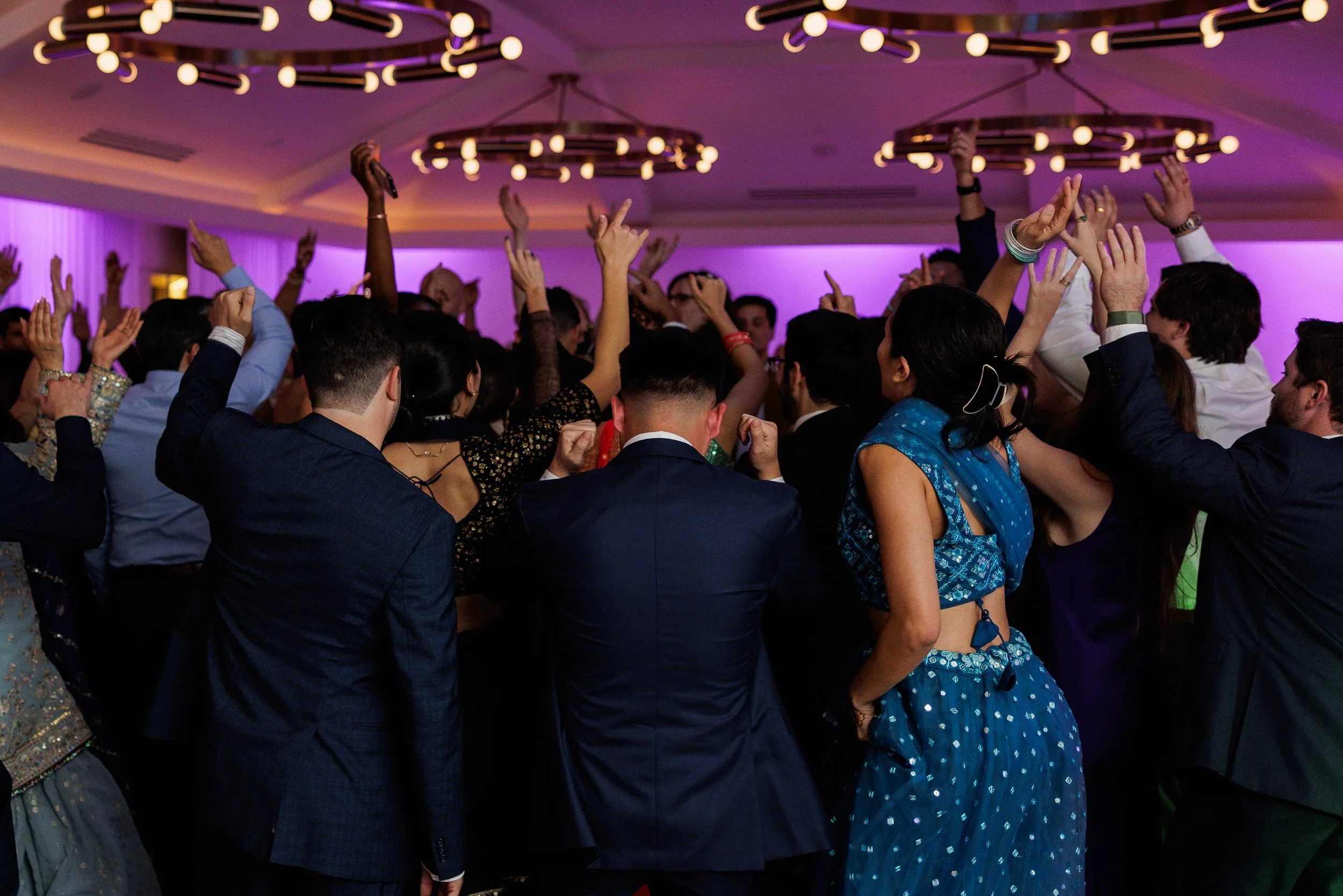 People dancing and celebrating at a party or wedding reception in a decorated indoor venue with purple lighting.
