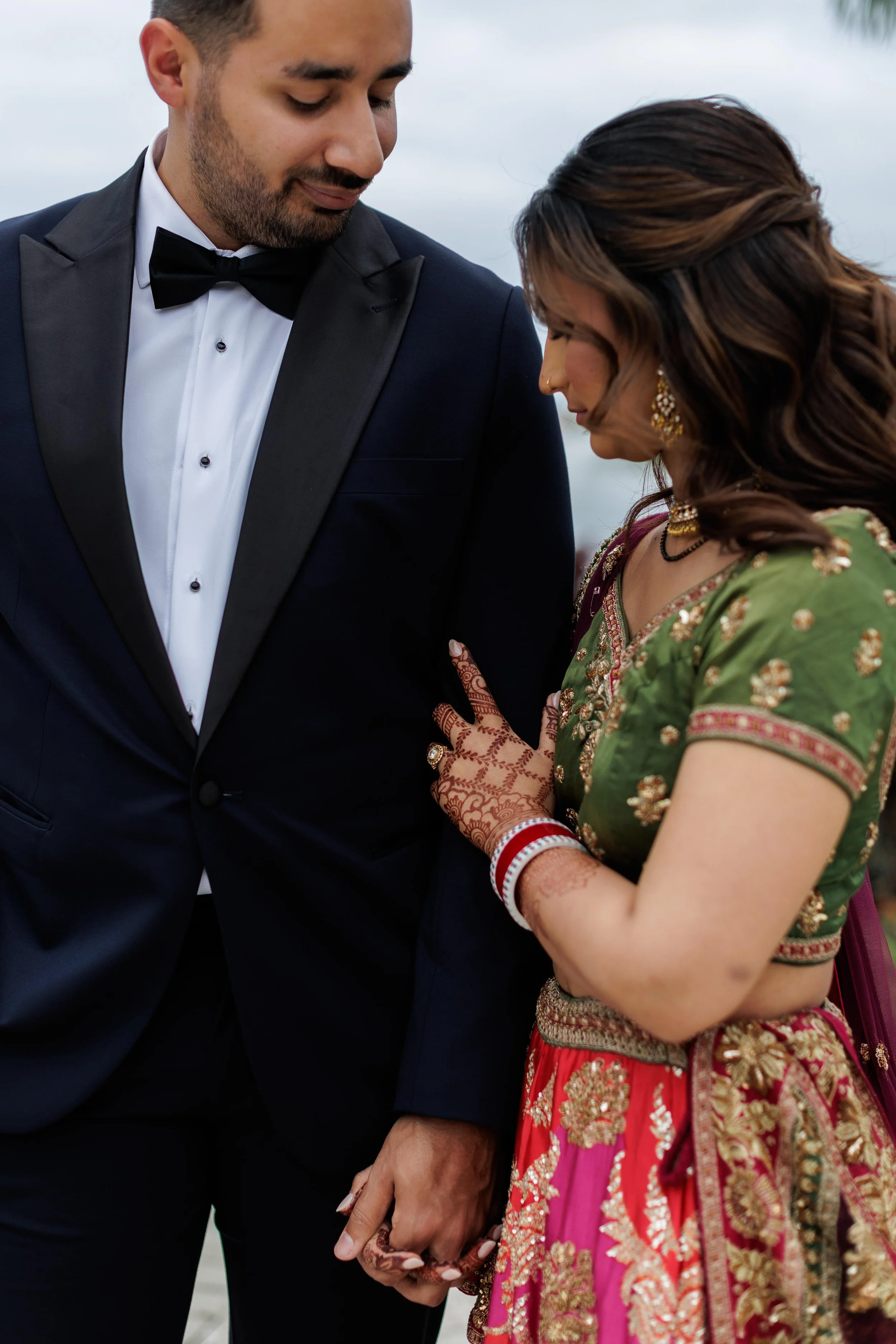 A man in a black tuxedo holding hands with a woman dressed in traditional Indian attire, the woman wearing henna on her hands, bangles, earrings, and a green and pink outfit, with their heads bowed and eyes closed.